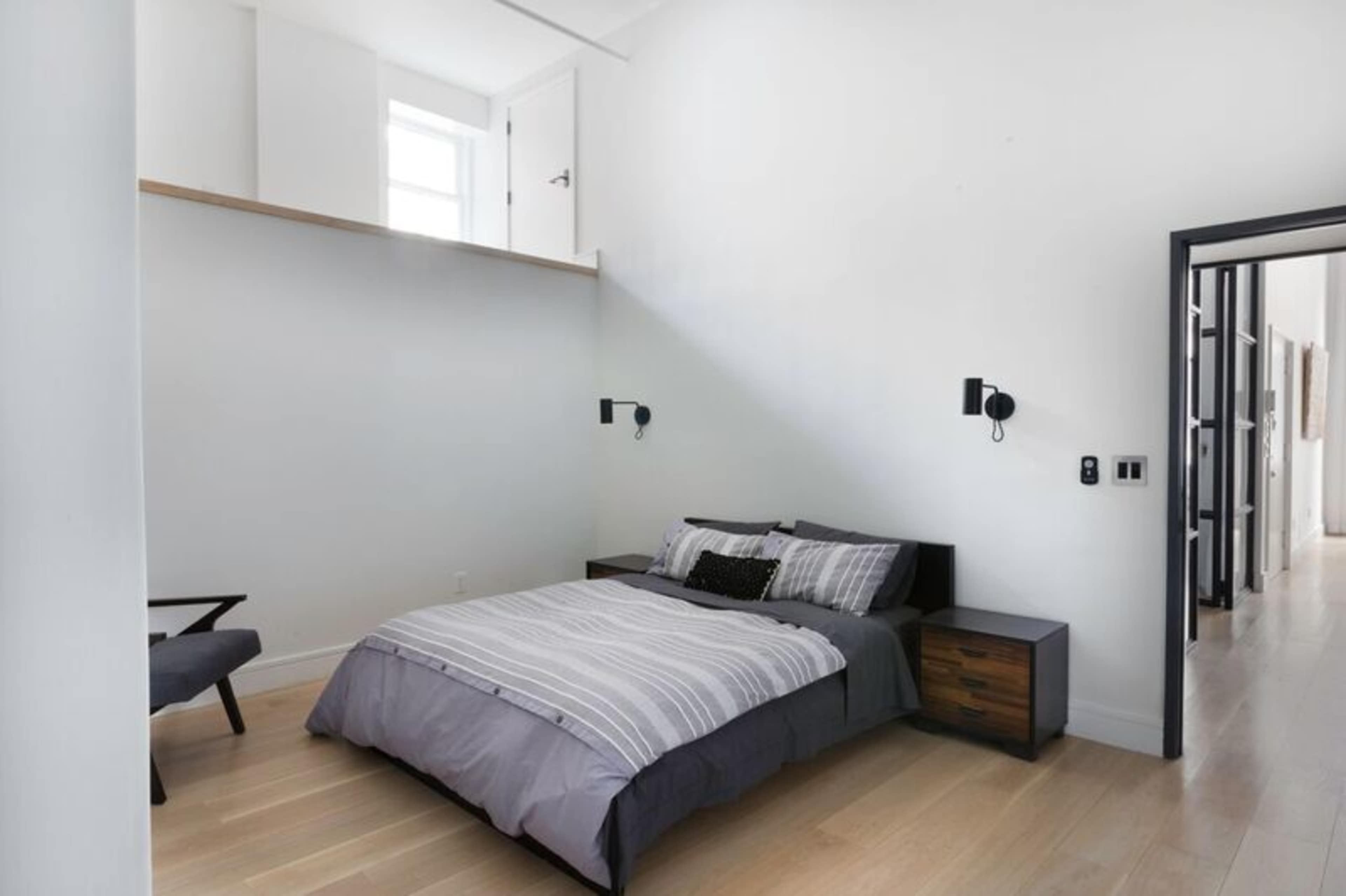 A minimalist bedroom with a bed, bedside tables, and a chair, featuring white walls and wooden flooring.
