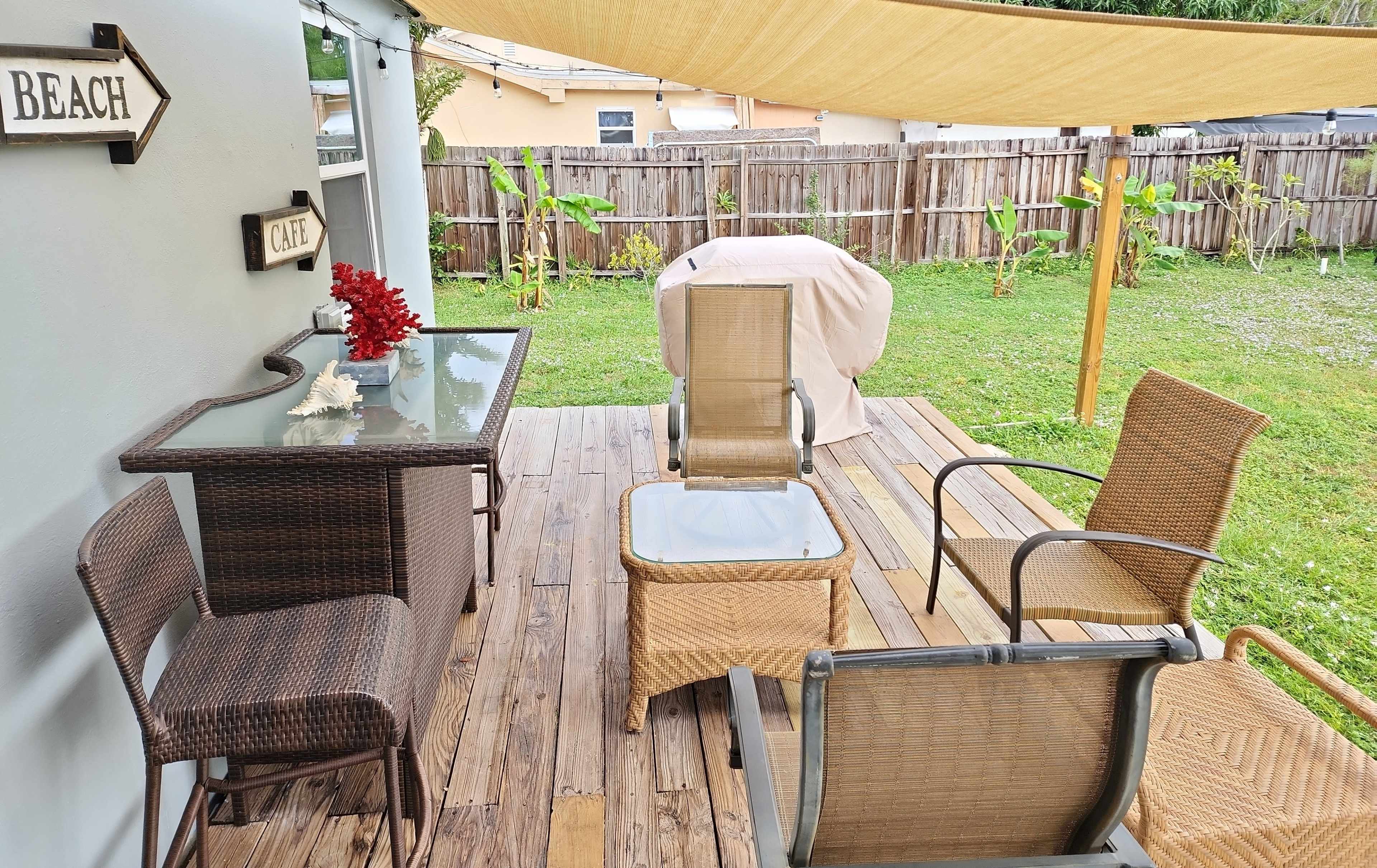 A patio area with wicker furniture arranged around a table, a grill covered with a cloth, and a slanted shade sail overhead, set against a grassy backdrop with a wooden fence.