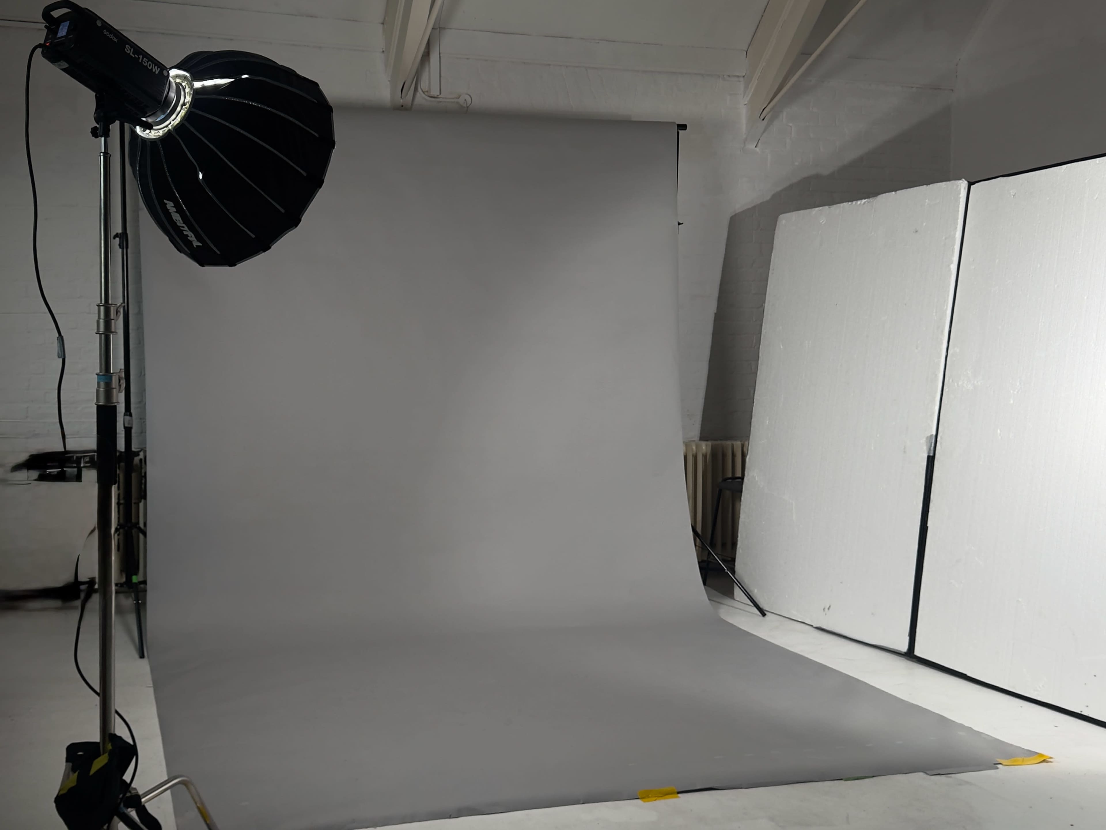 The image shows a photography studio with a gray backdrop, a light source with a softbox, and two large white reflectors positioned nearby.