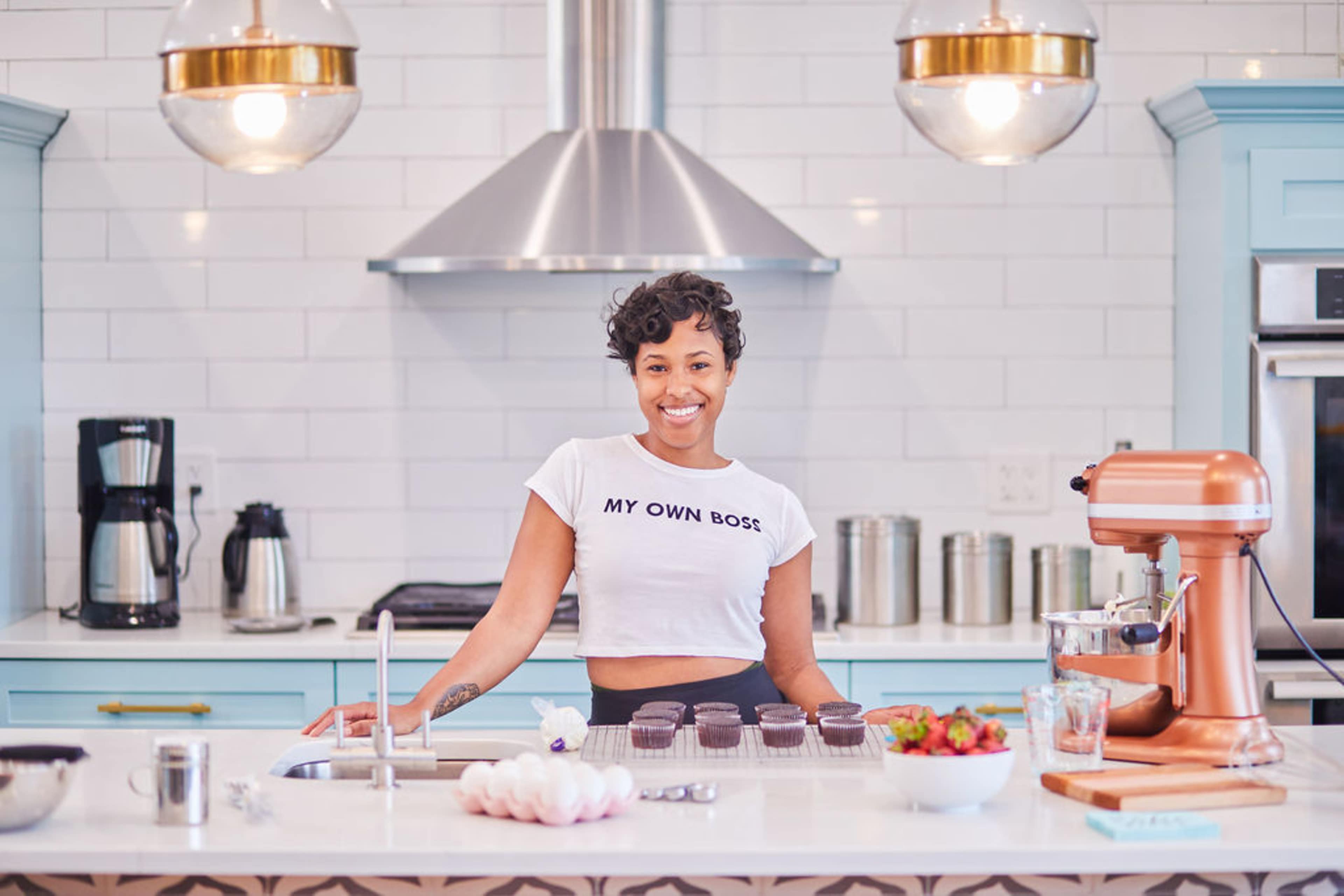 A woman stands in a modern kitchen, smiling while wearing a shirt that says "MY OWN BOSS" and surrounded by baking ingredients and equipment.