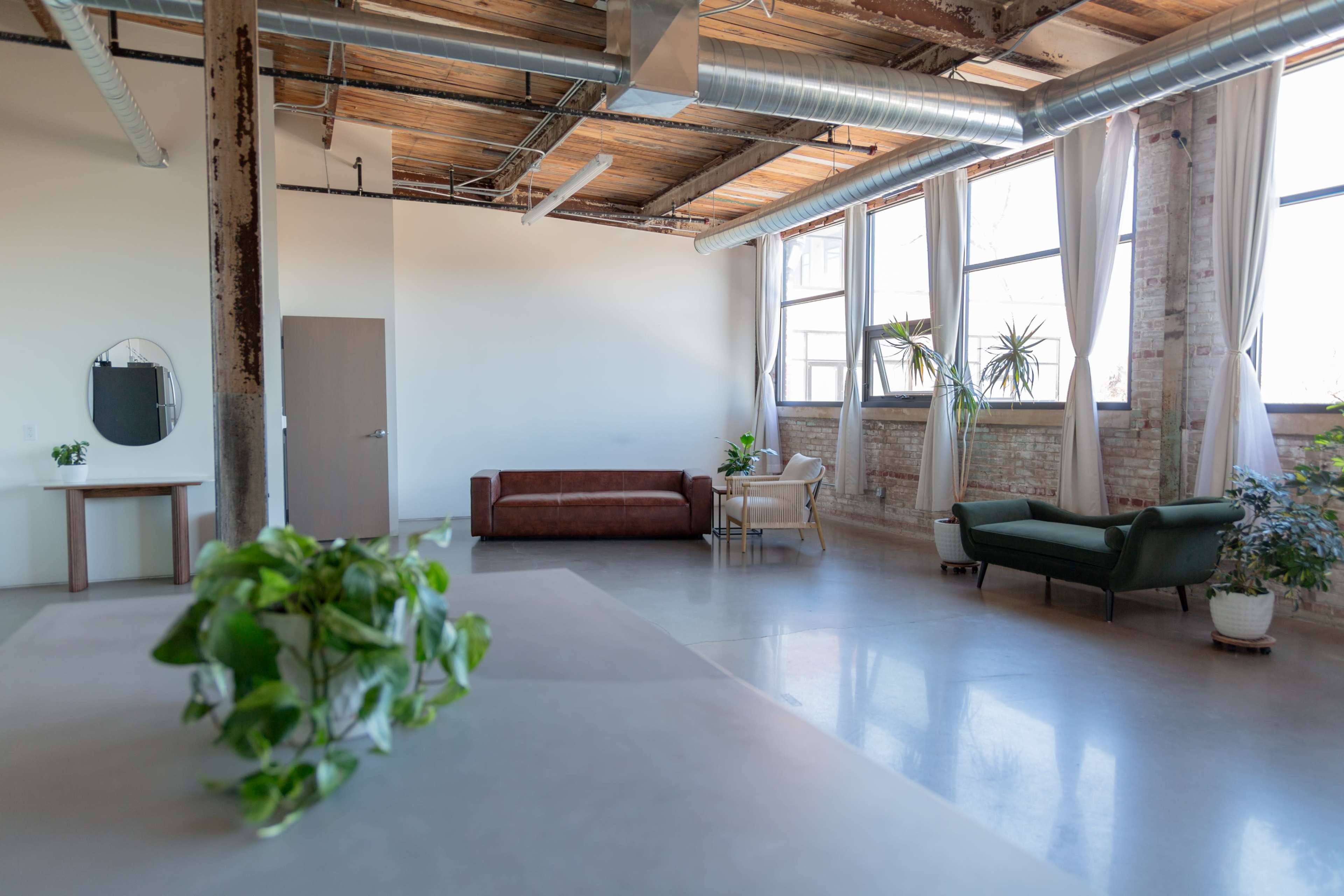 The image shows a spacious, modern interior with exposed beams, large windows, and a mix of furniture, including a brown couch and a green chair.