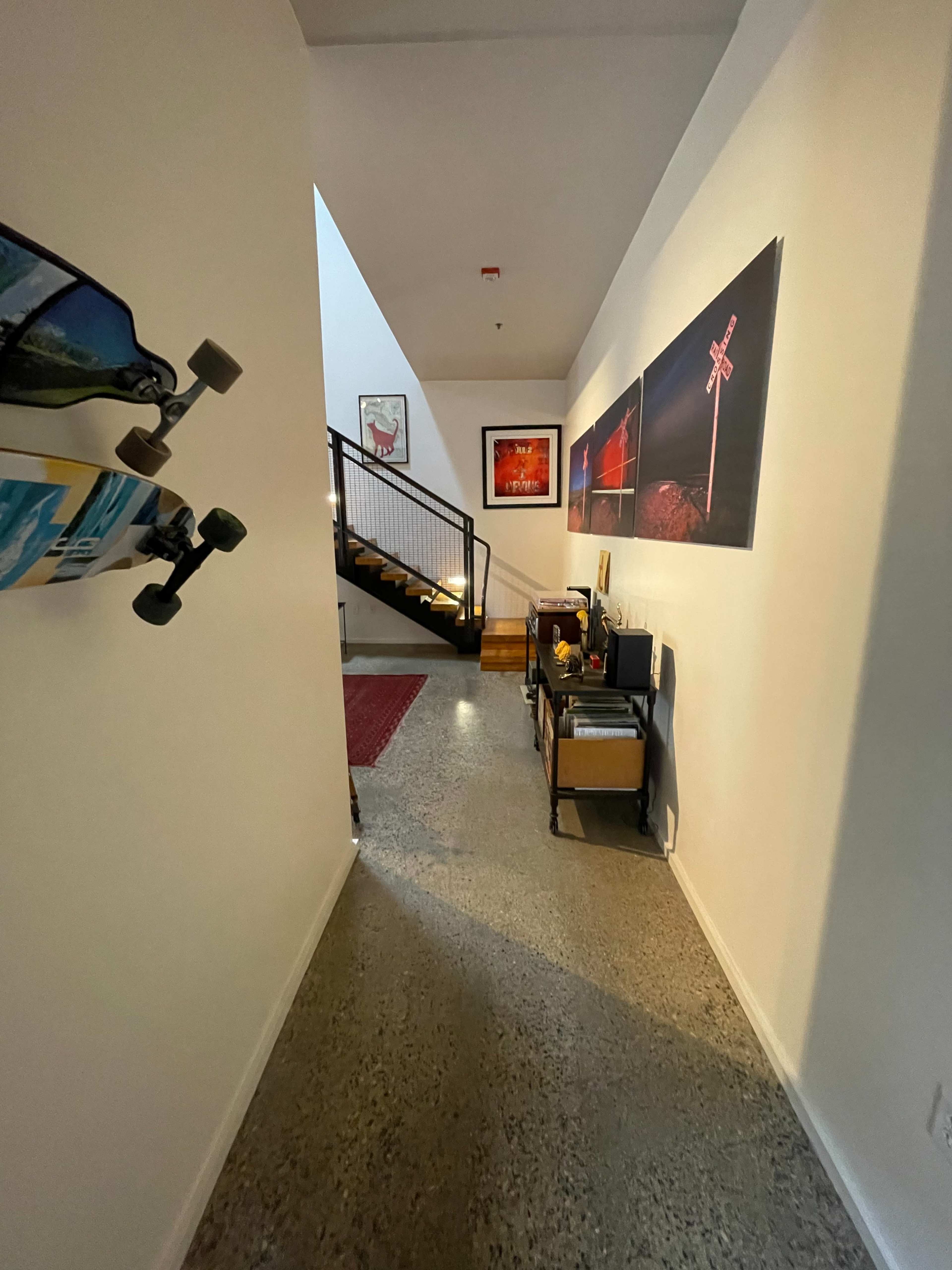 A narrow hallway leading to a staircase, with a skateboard mounted on the wall and framed photographs hanging on the opposite wall.