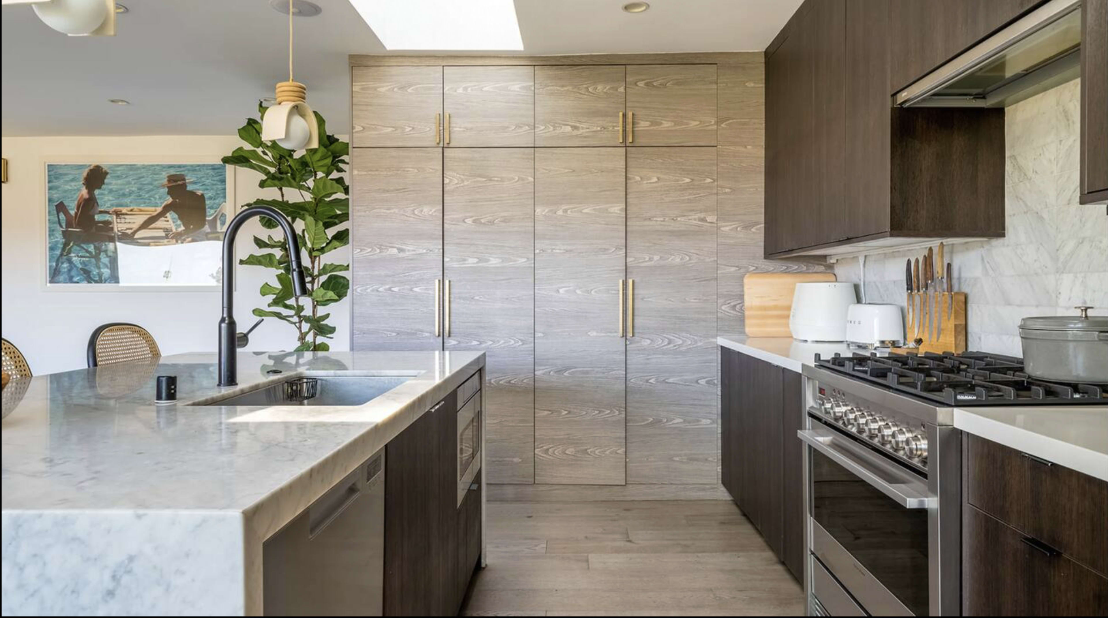 A modern kitchen features a marble island, wooden cabinetry, and stainless steel appliances, with a large window allowing natural light.