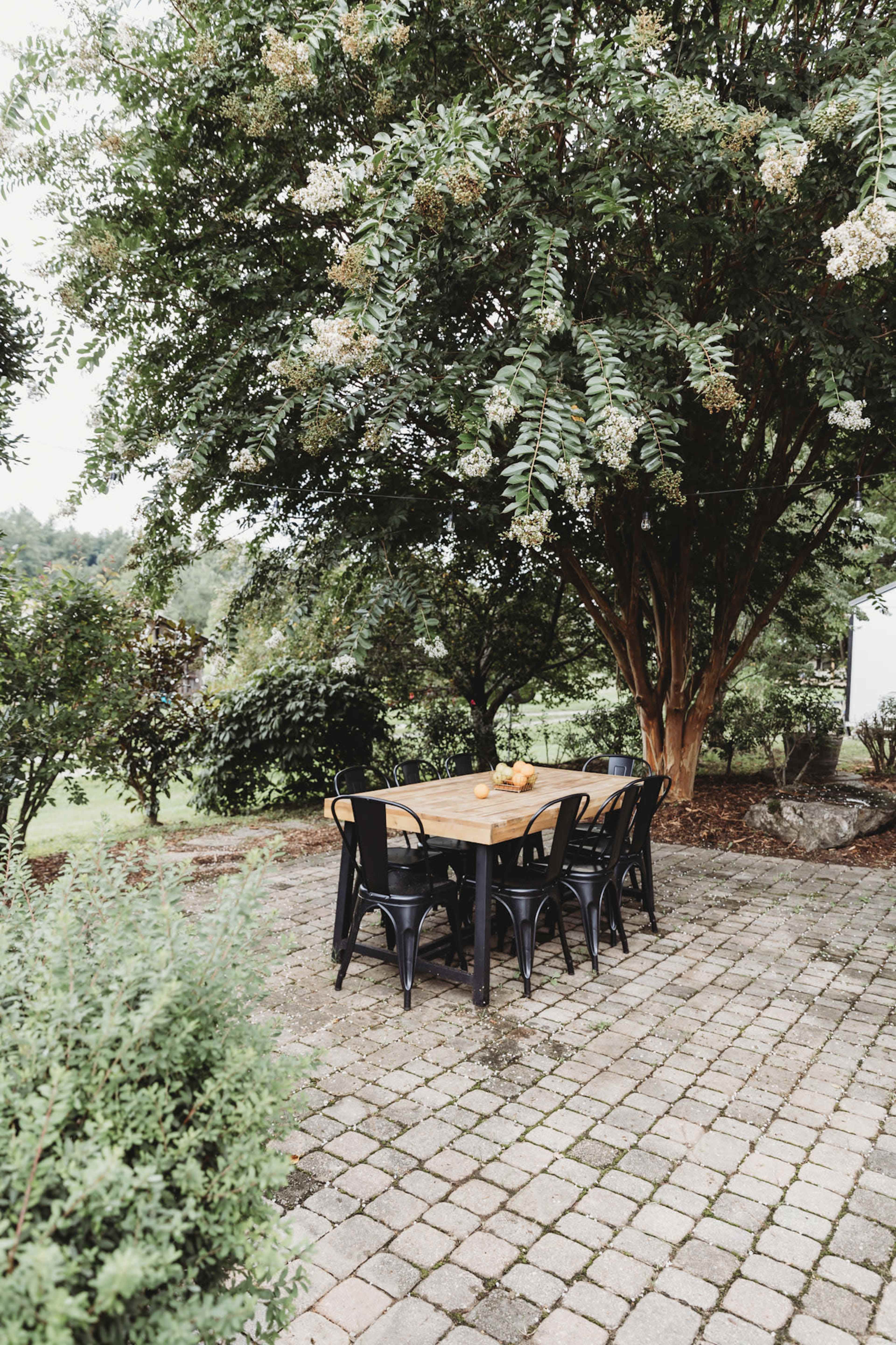 A rectangular wooden dining table with black chairs is set on a stone patio under a large tree.
