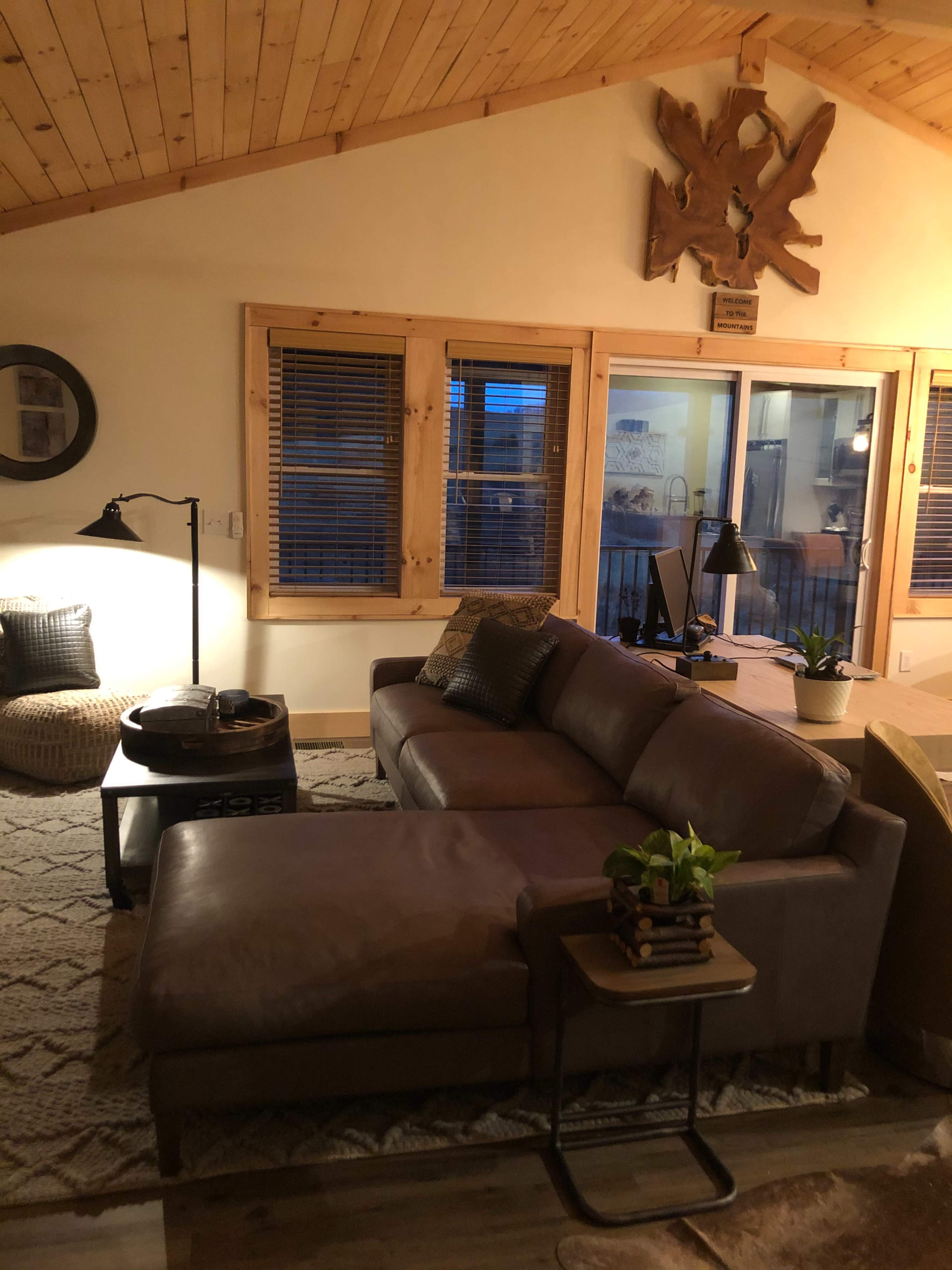 A cozy living room with a leather sectional sofa, a round coffee table, a floor lamp, and large windows that allow natural light to enter.