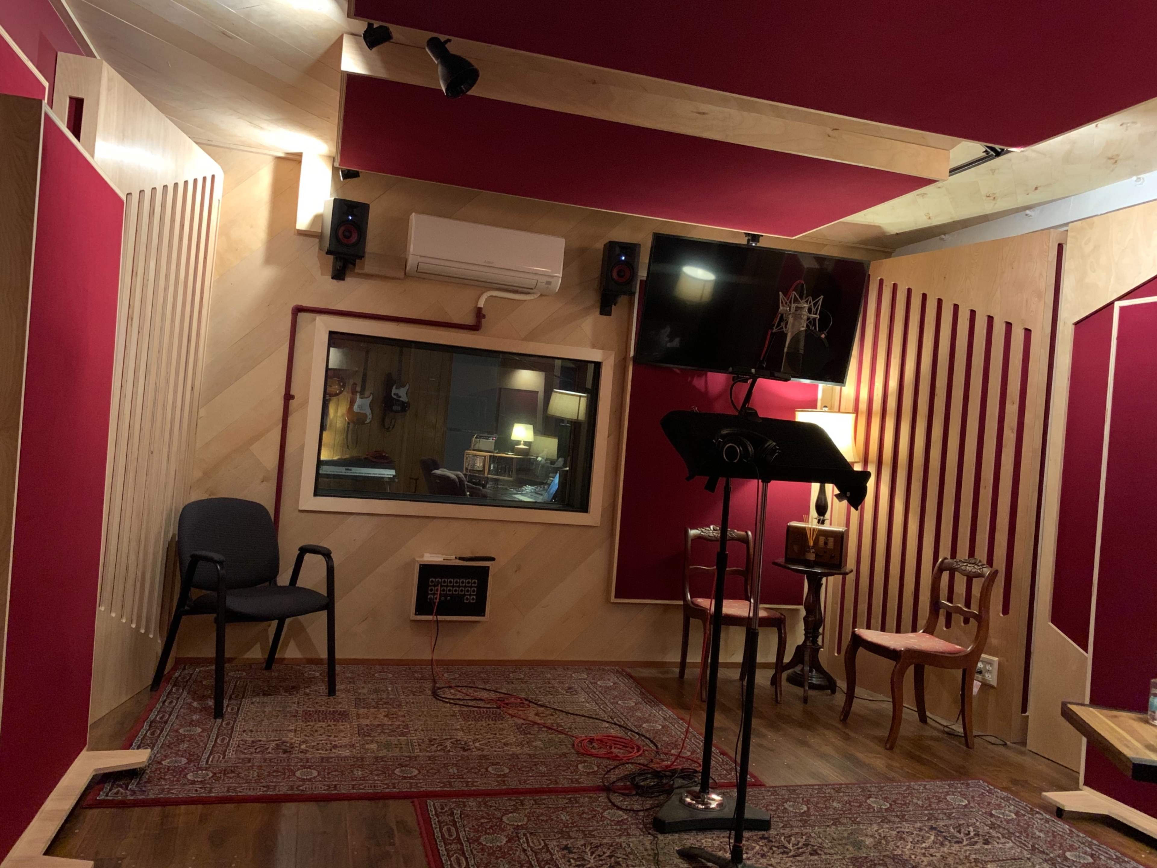A recording studio with soundproofed walls, a control room visible through a window, and various seating arrangements including a chair and a stand for scripts.