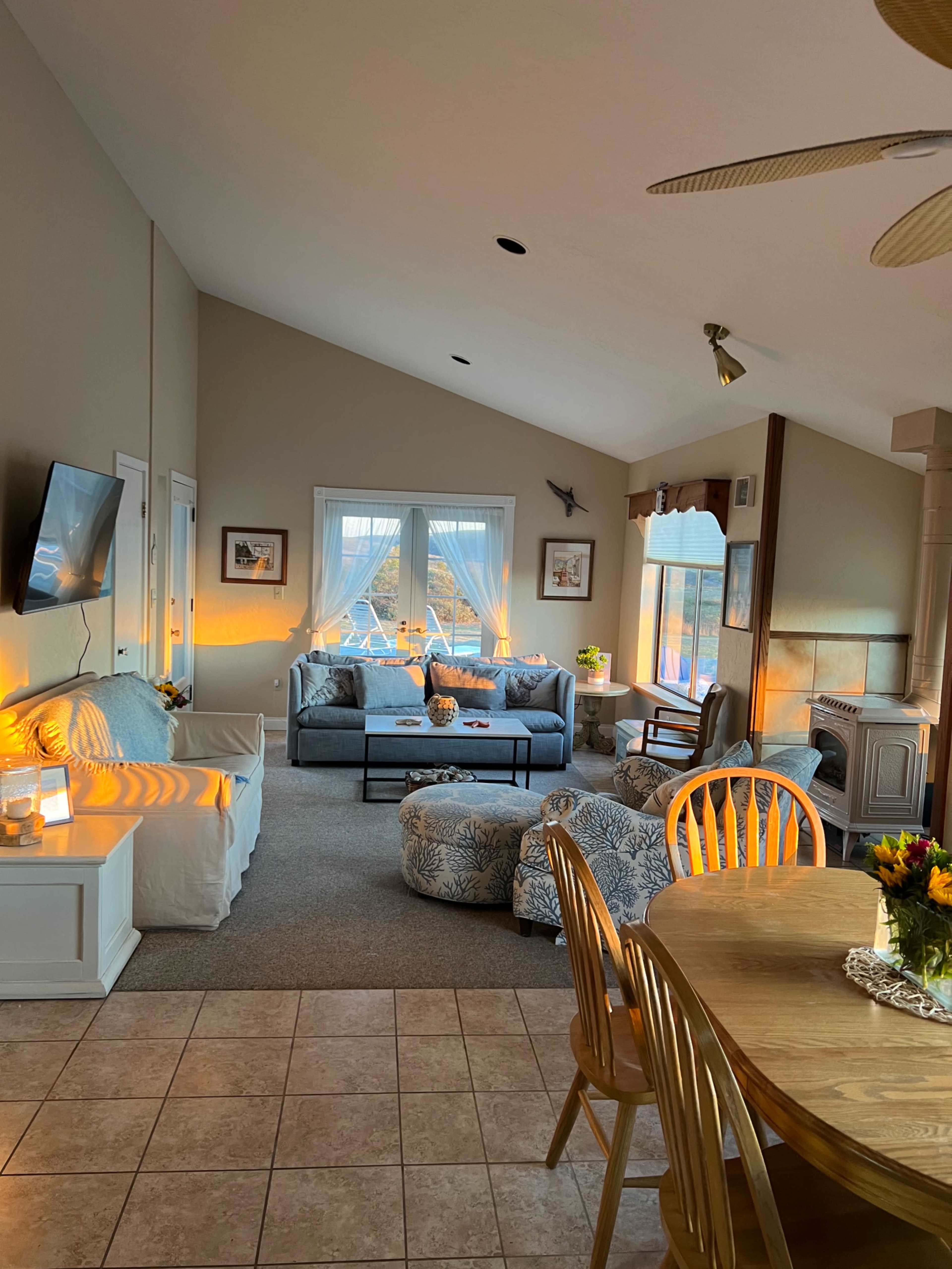 A spacious living area features a seating arrangement with a sofa and armchairs, a dining table, and large windows allowing natural light to fill the space.