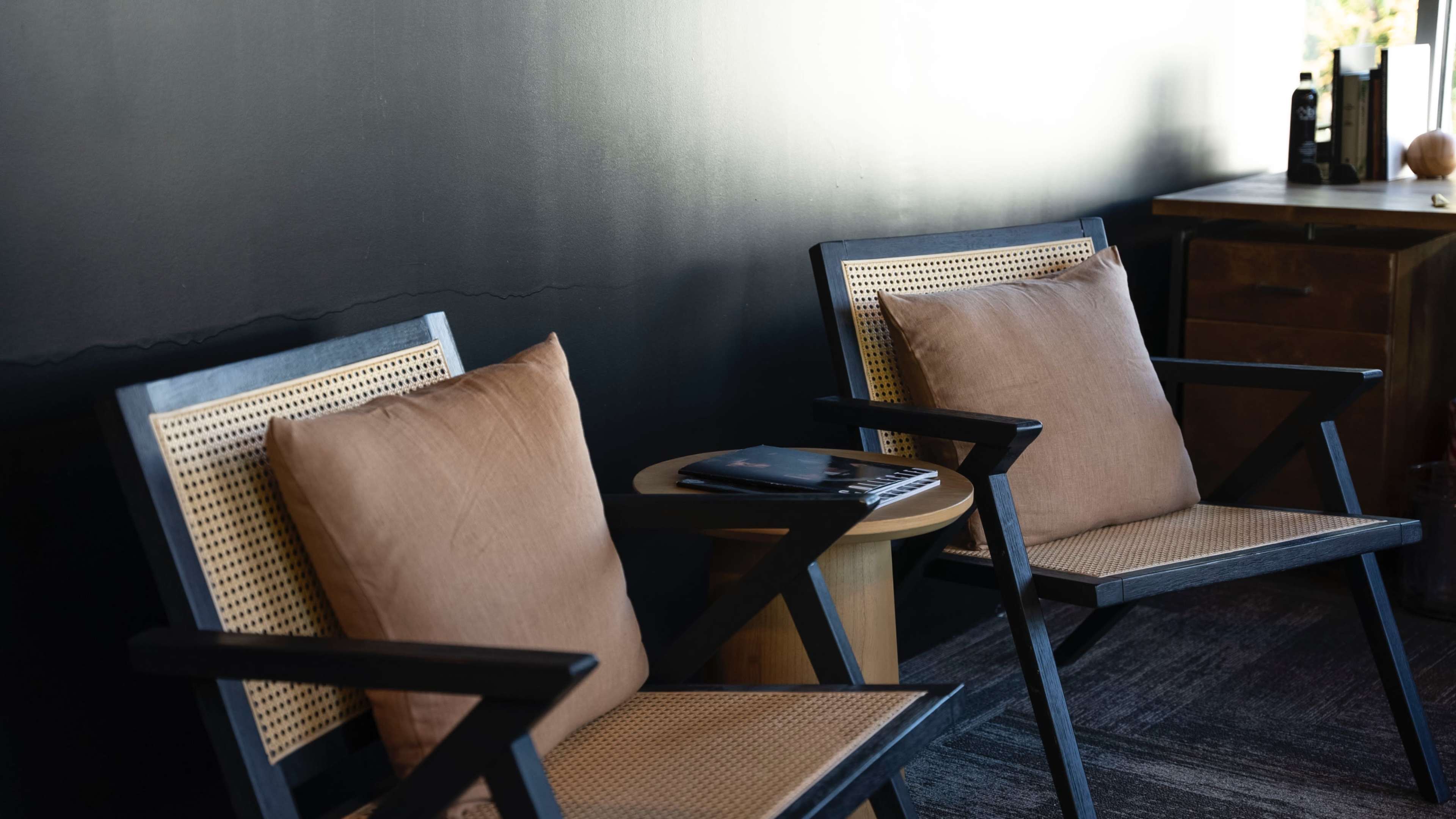 Two wooden chairs with cushions sit beside a small round table against a black wall.