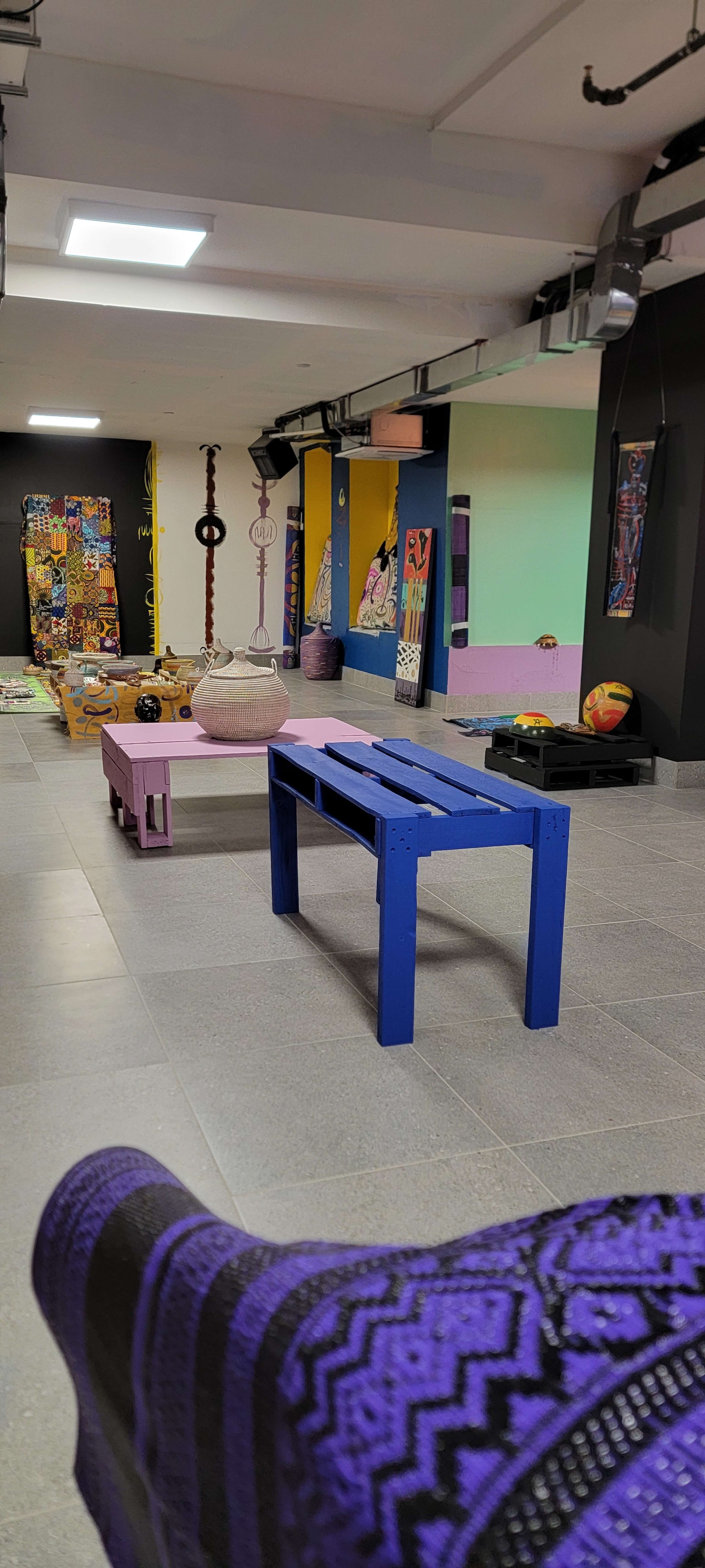 The image shows an indoor space with colorful walls, various art pieces displayed on the floor, and a table painted blue in the center.