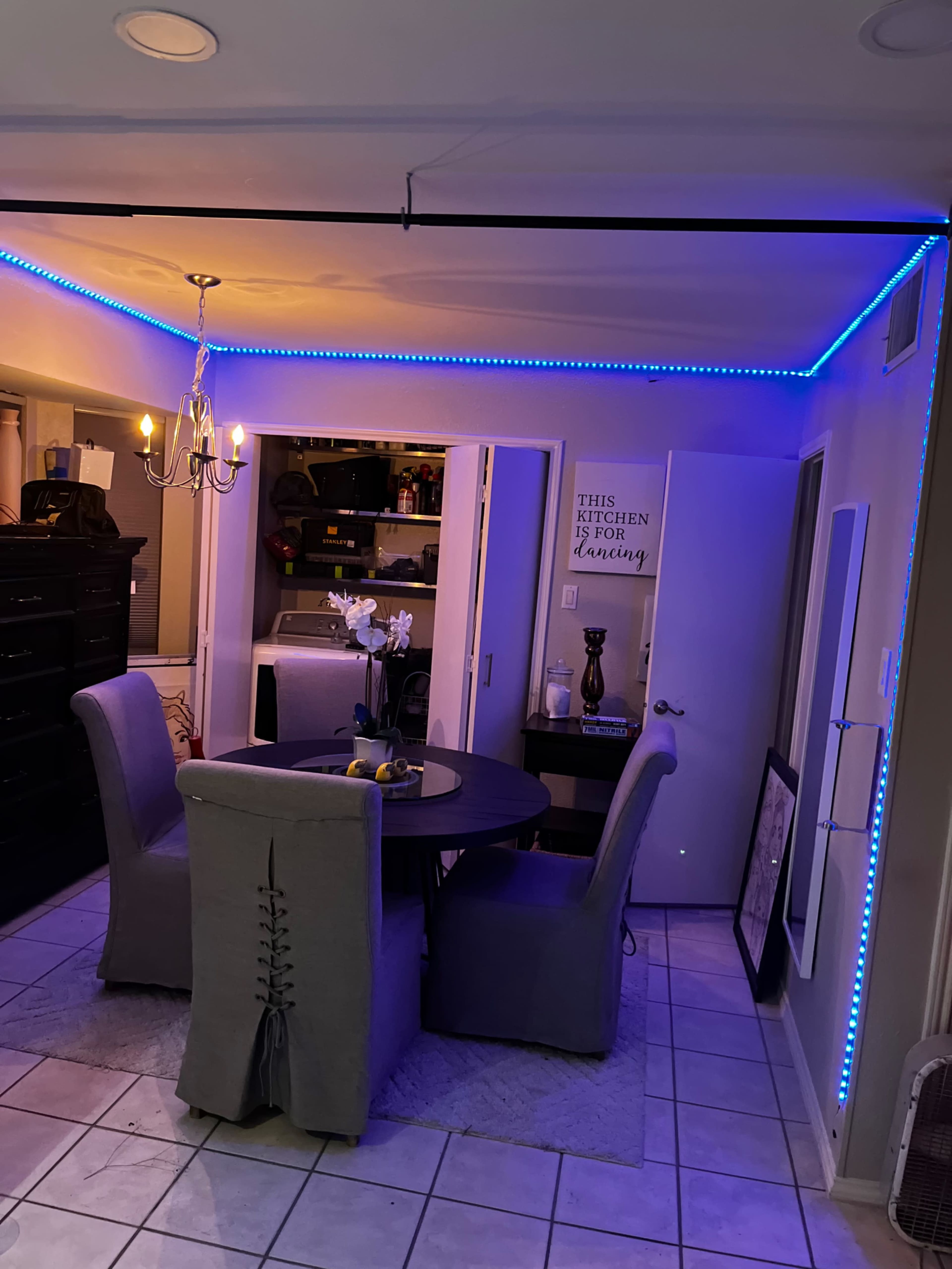 A round dining table with four upholstered chairs is illuminated by blue LED strip lighting in a room featuring a chandelier and a wall with decorative signs.