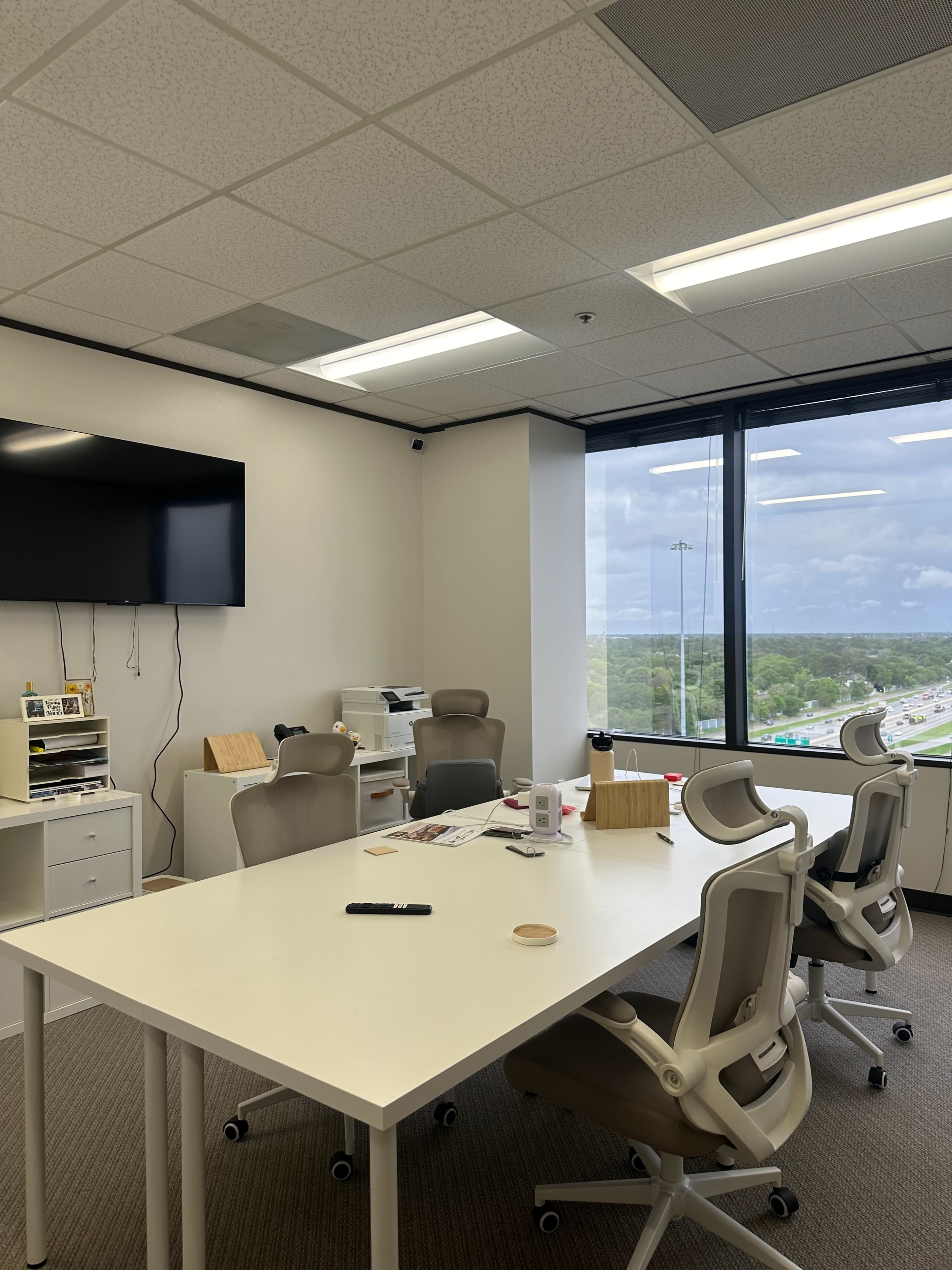 A well-lit office meeting room features a large white table, several ergonomic chairs, a wall-mounted television, and a view of the highway through large windows.