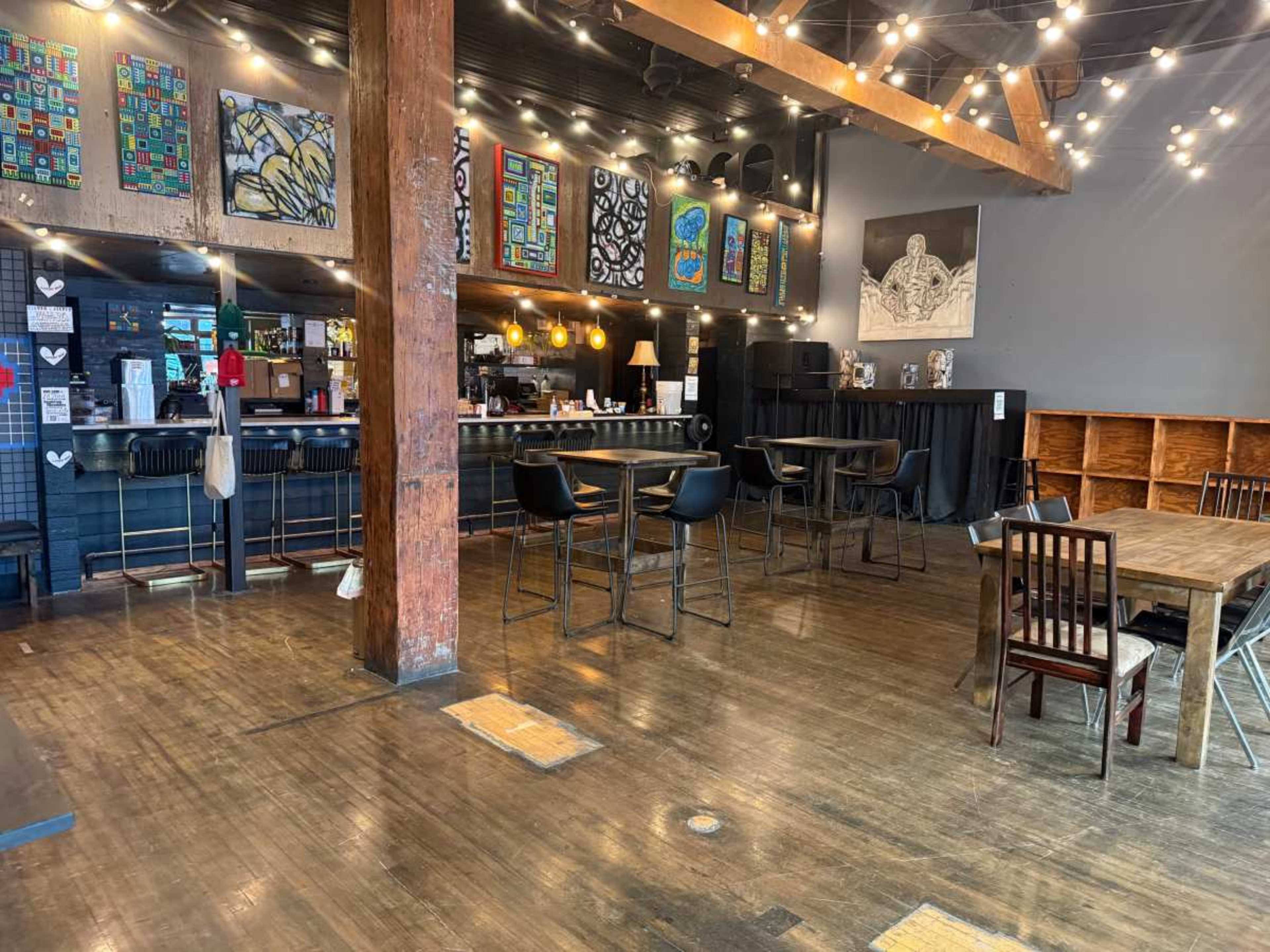 The image shows a spacious café with wooden floors, a bar area in the background, and various artwork displayed on the walls.