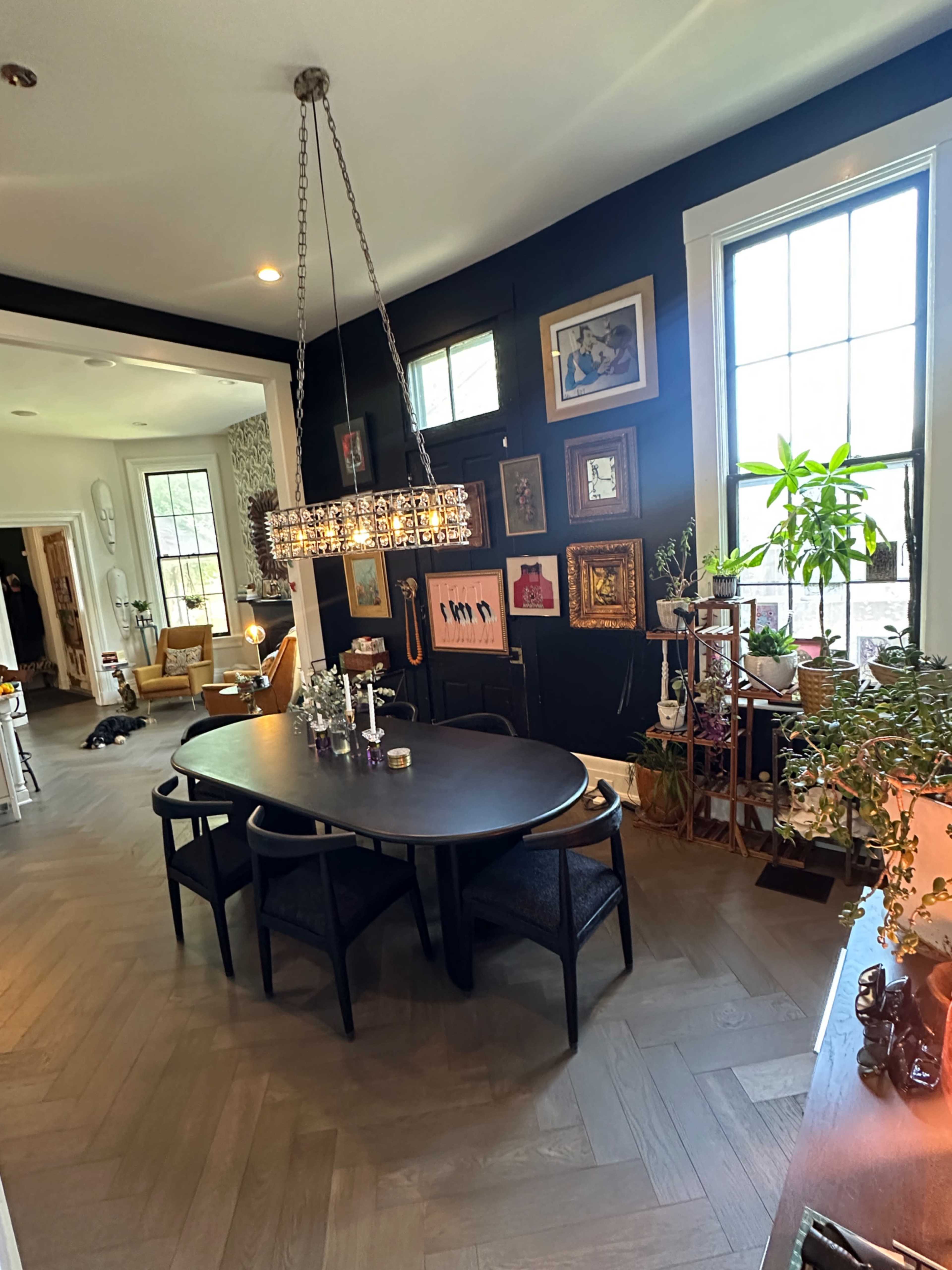 The image shows a dining area featuring a dark table surrounded by chairs, a decorative chandelier, and walls adorned with various framed artworks.
