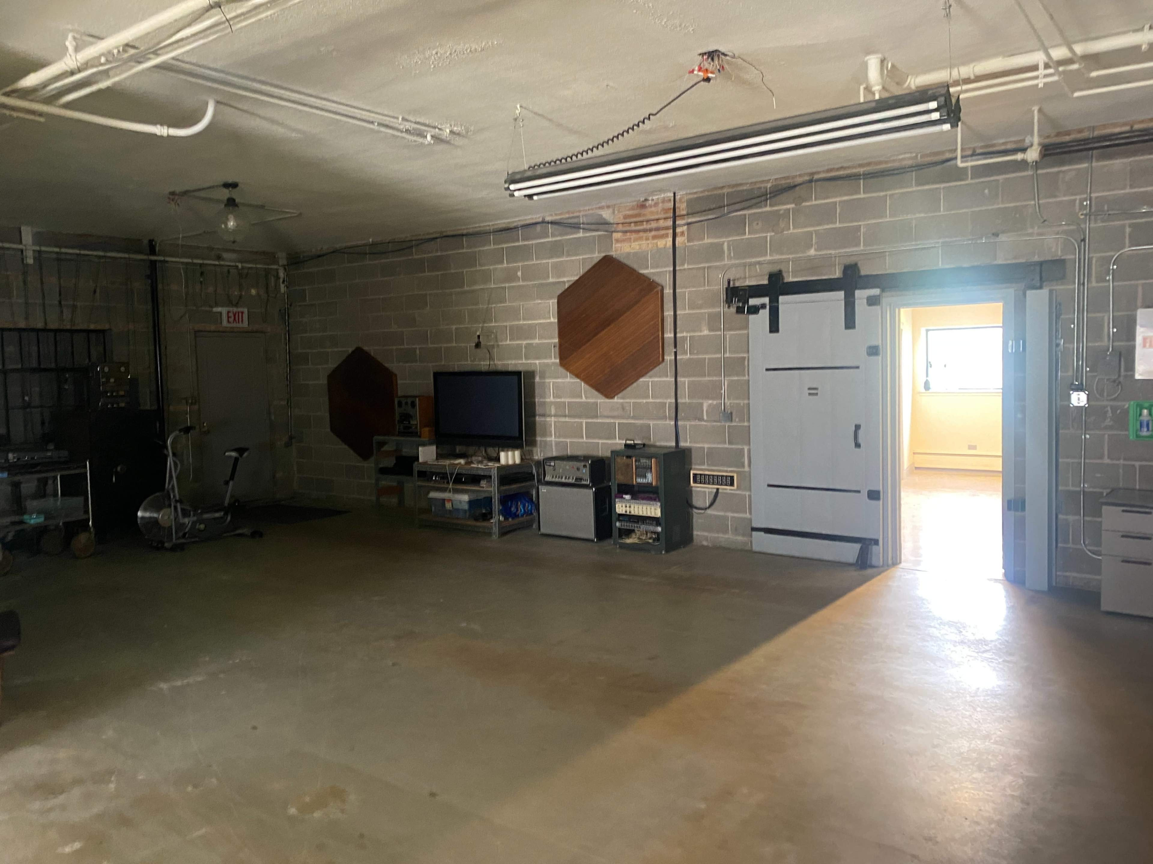 The image shows an empty industrial-style room with concrete walls, a television on a stand, and a doorway leading to another space.