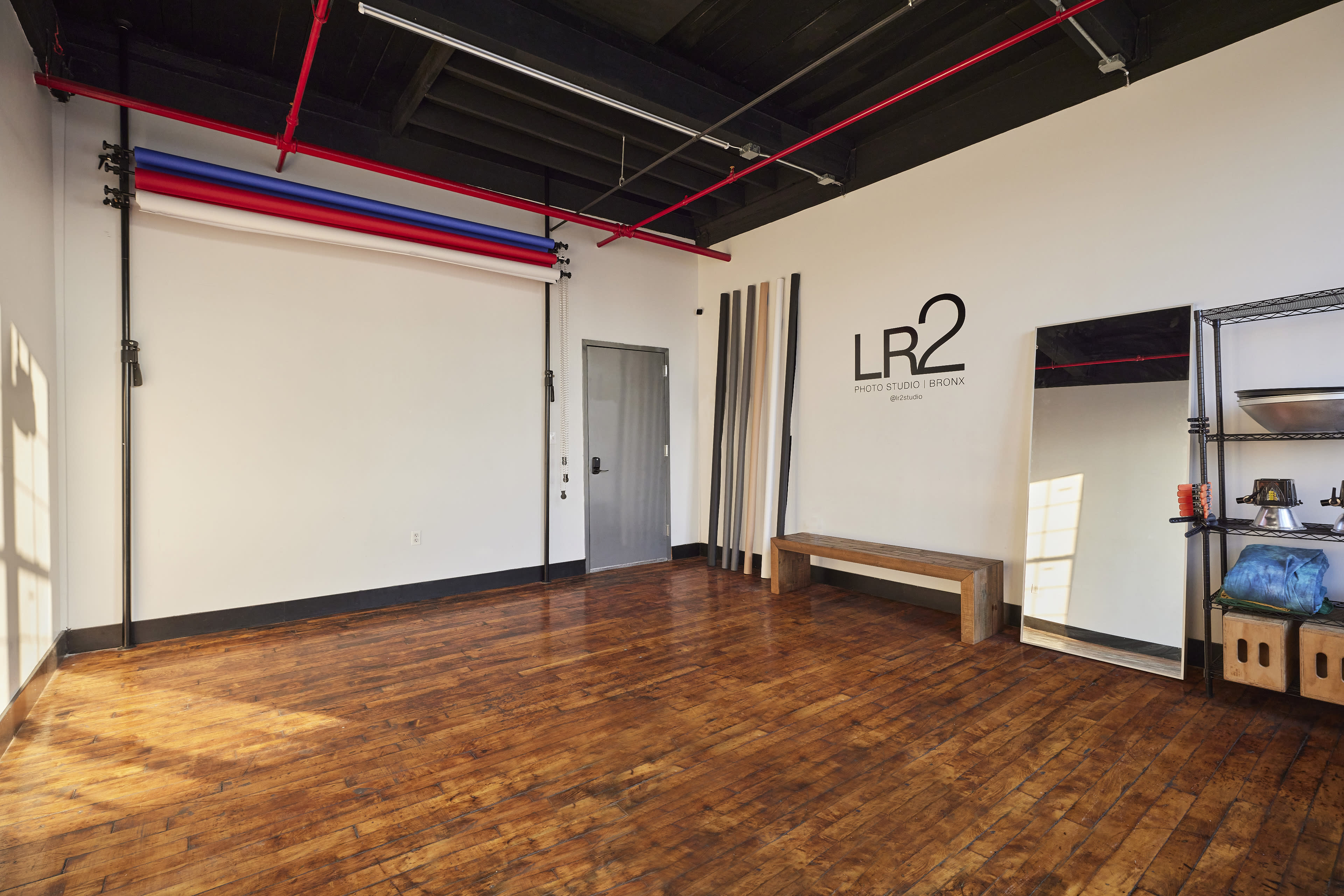 A spacious, empty studio with wooden flooring, a mirrored wall, a bench, and a few vertical props against a white backdrop.
