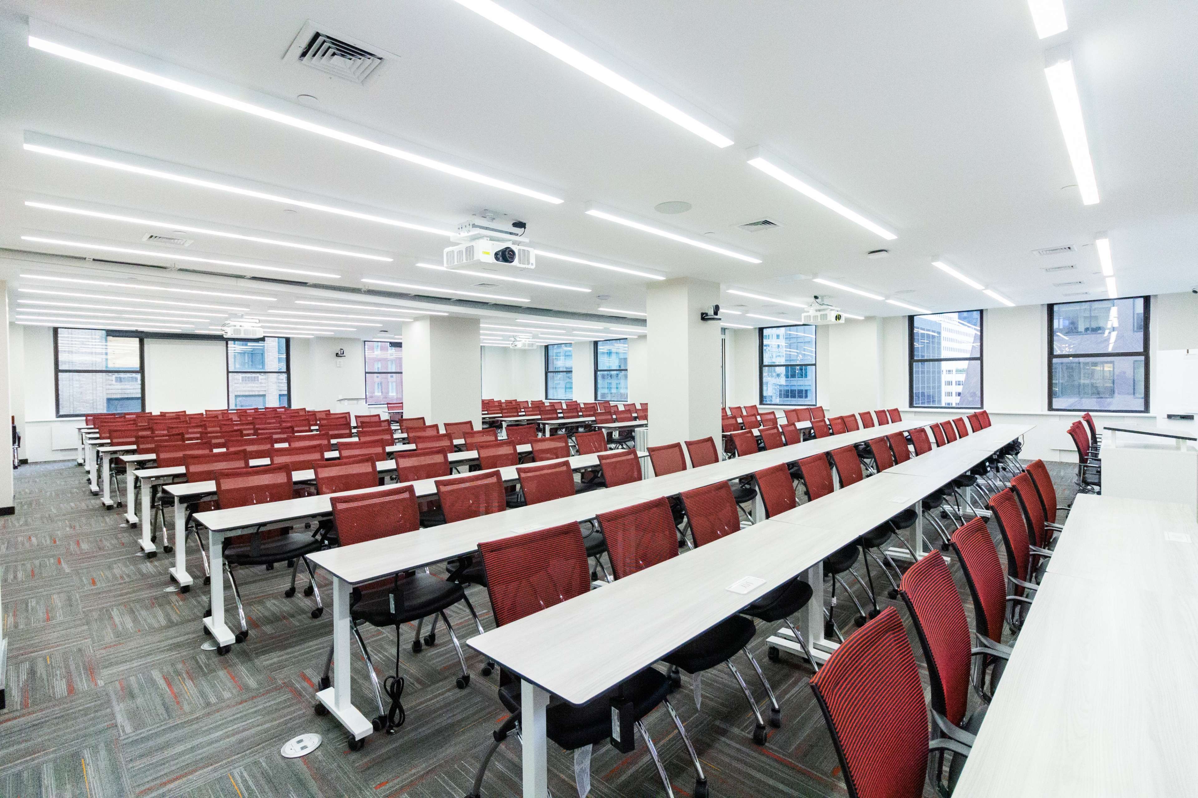 The image shows a modern, spacious classroom with rows of tables and red chairs arranged for a seminar or meeting.