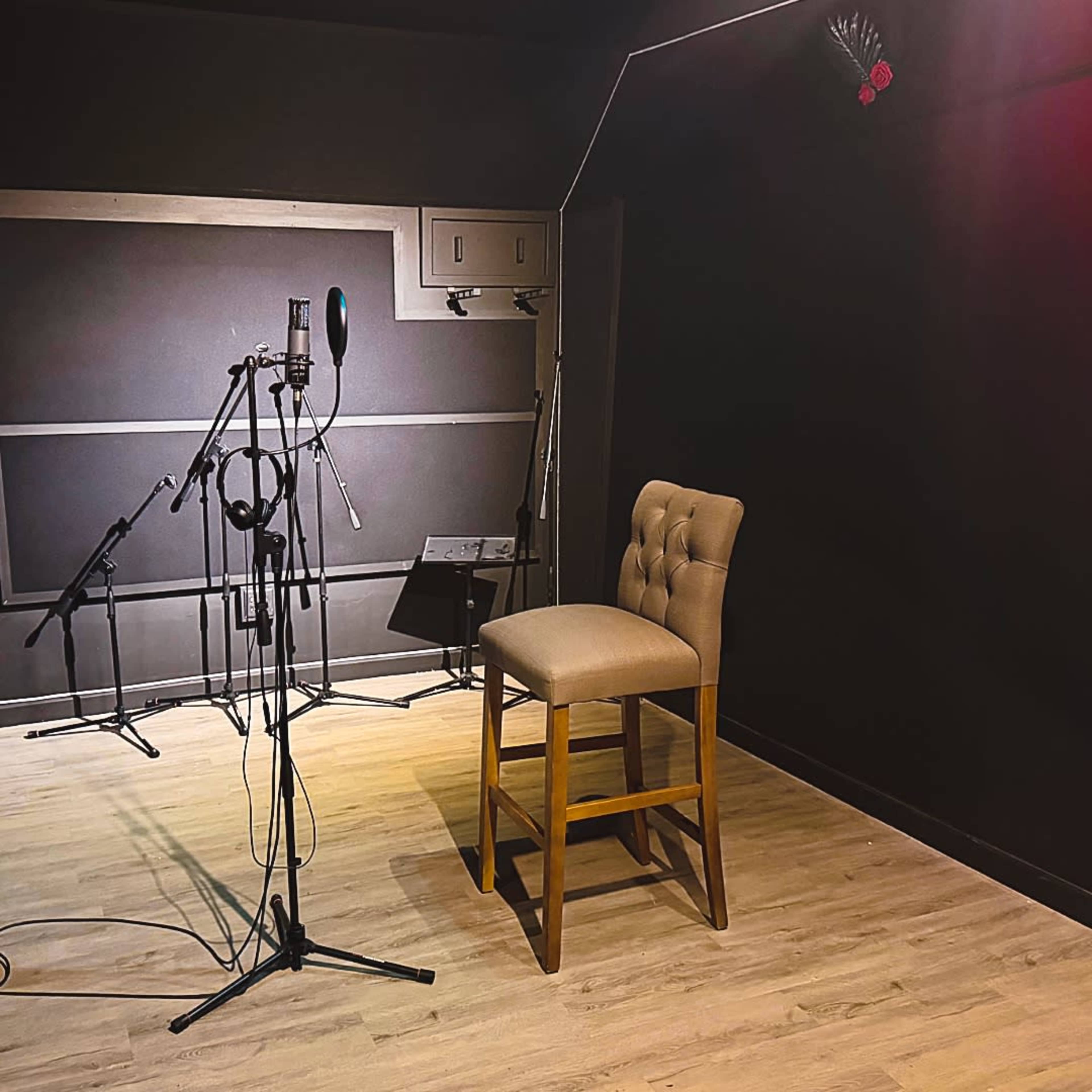 The image shows a small recording studio with a single wooden stool, microphones, and headphones arranged in front of a dark wall.
