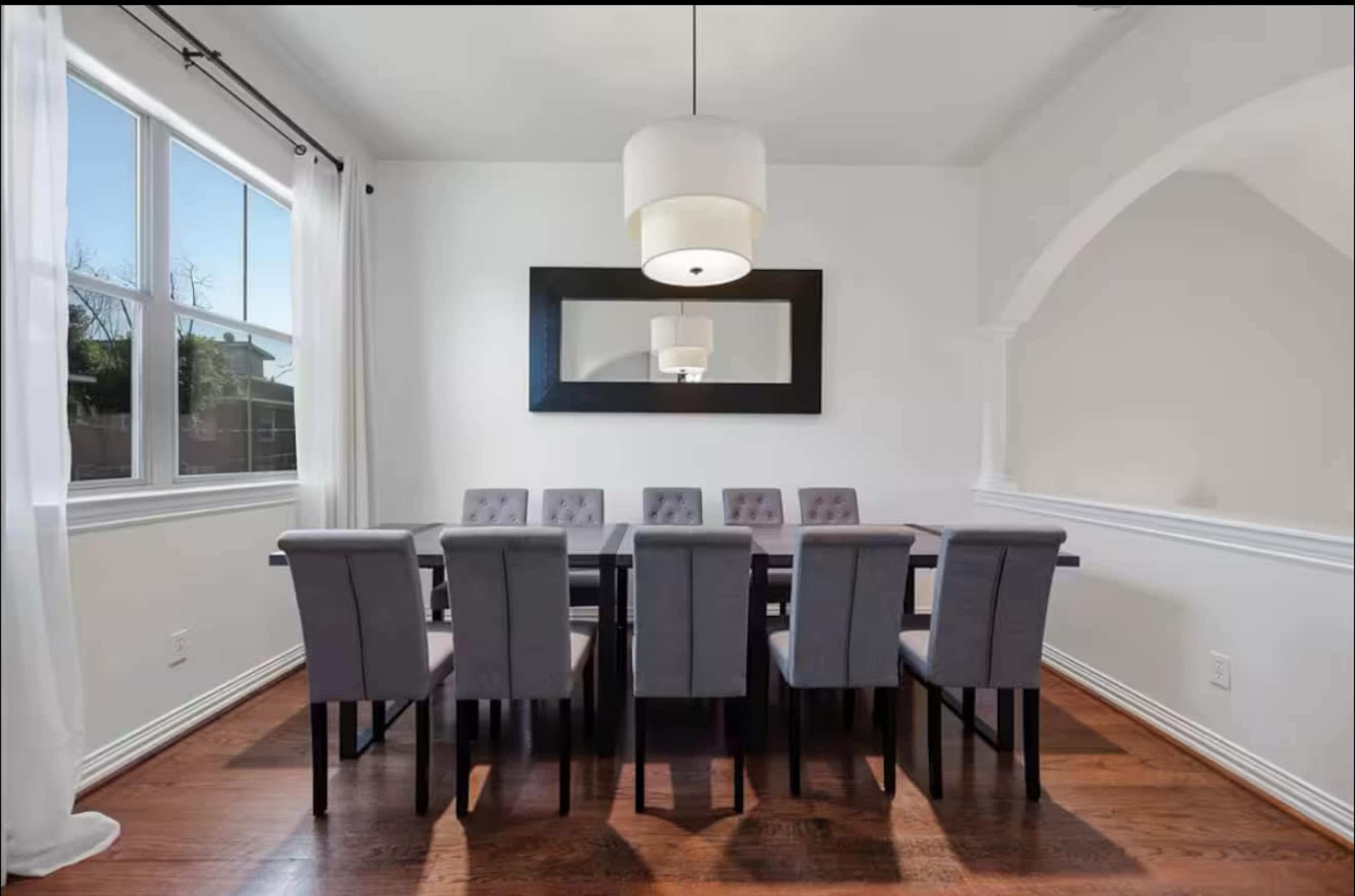 A large dining room features a rectangular table surrounded by eight upholstered chairs, with a wall mirror and a modern pendant light overhead.