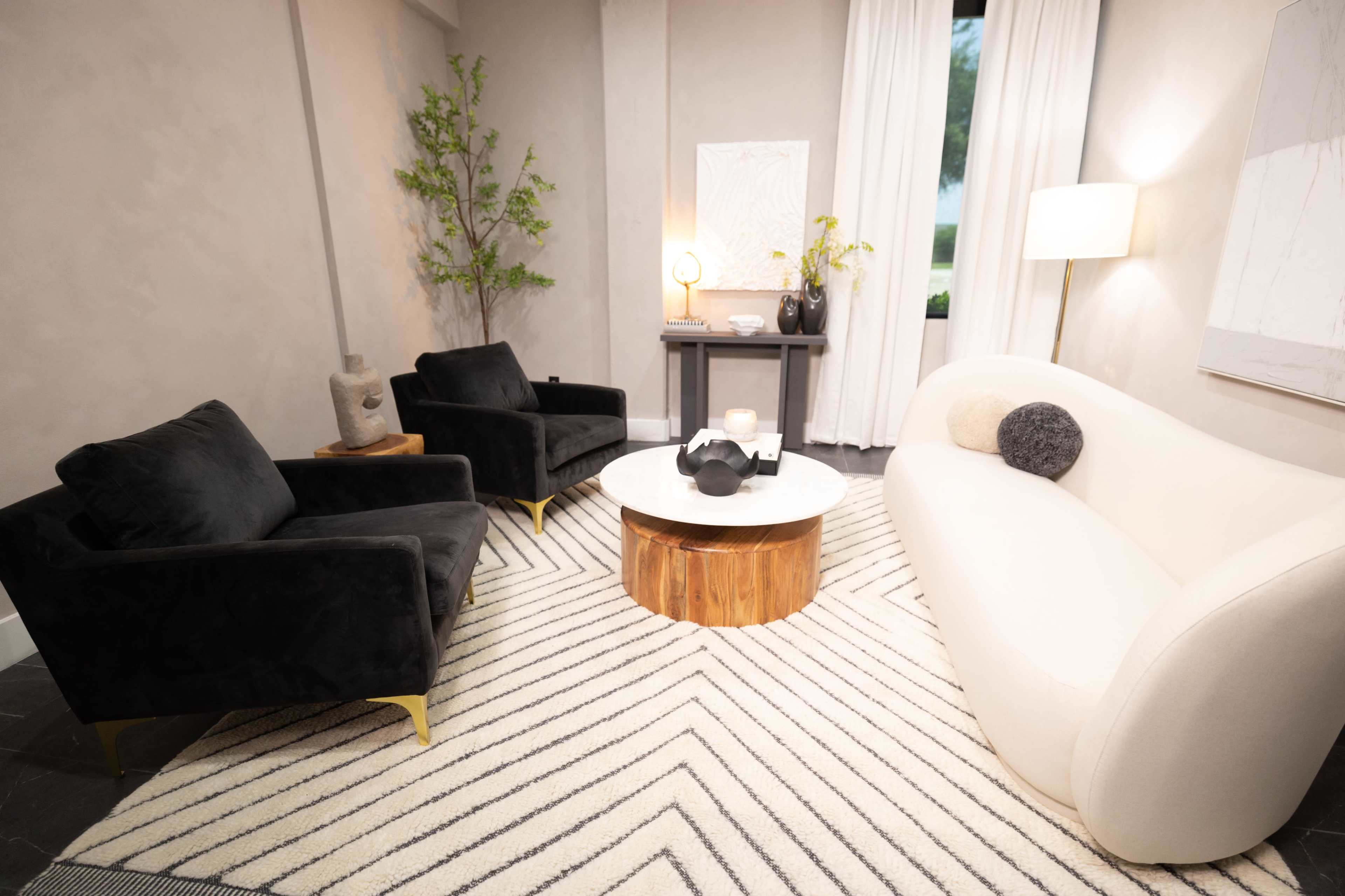 A modern living room setting with two black chairs, a white curved sofa, a wooden coffee table, and decorative elements against a neutral wall.