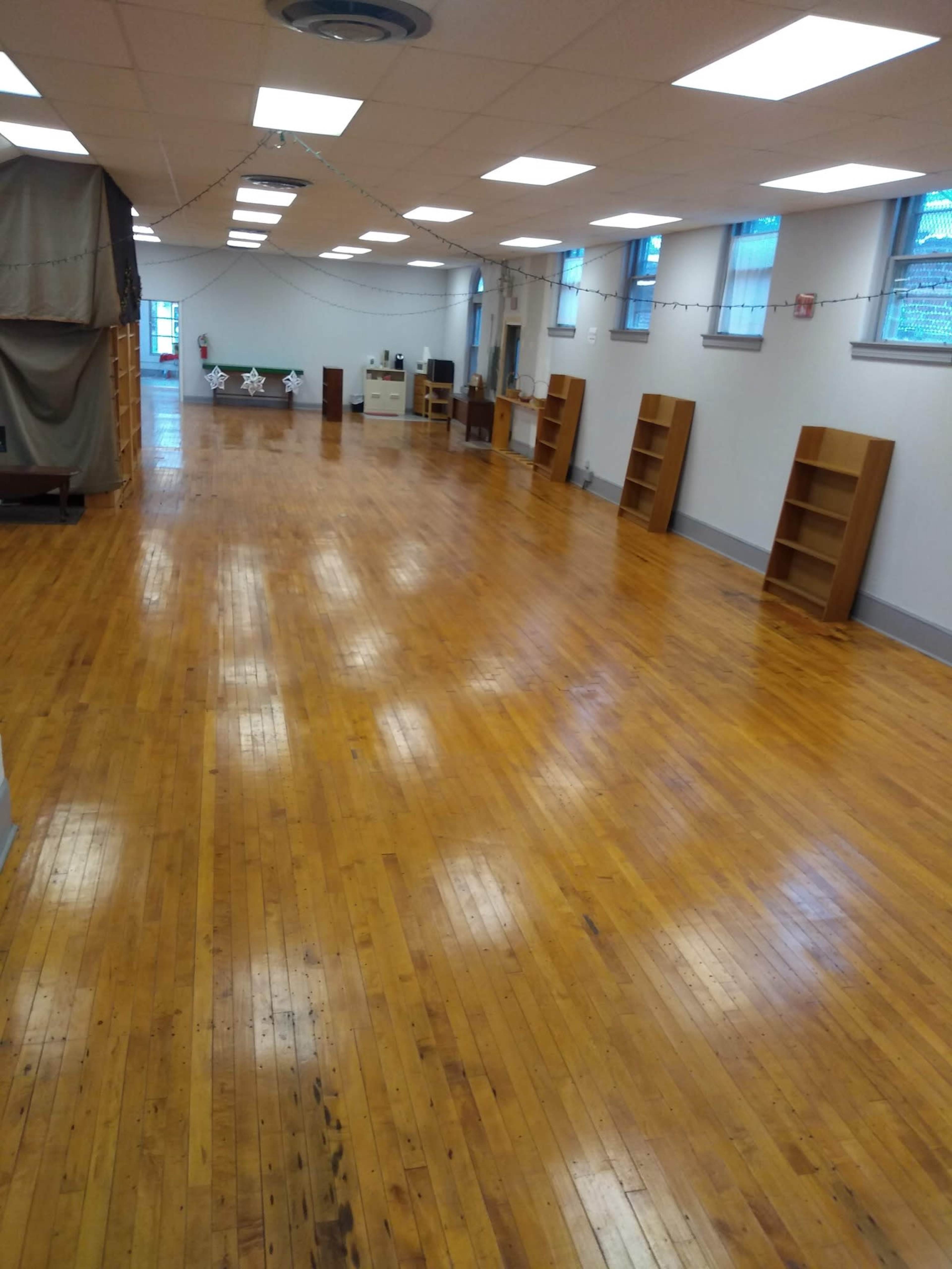 The image shows a spacious room with wooden floors, several empty bookshelves against the walls, and overhead fluorescent lighting.
