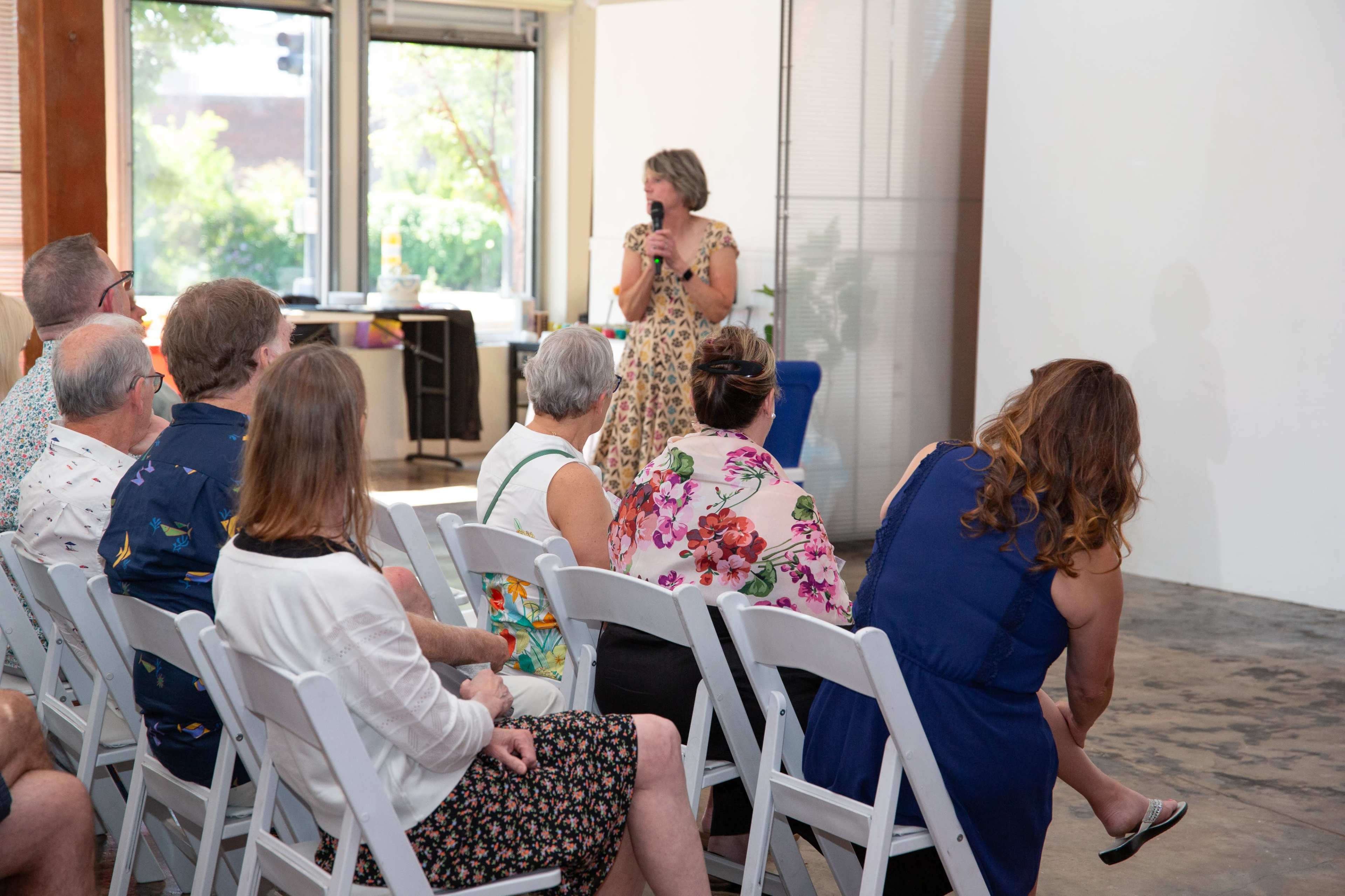 A woman stands at a microphone addressing an audience seated in white folding chairs in a bright, open space.