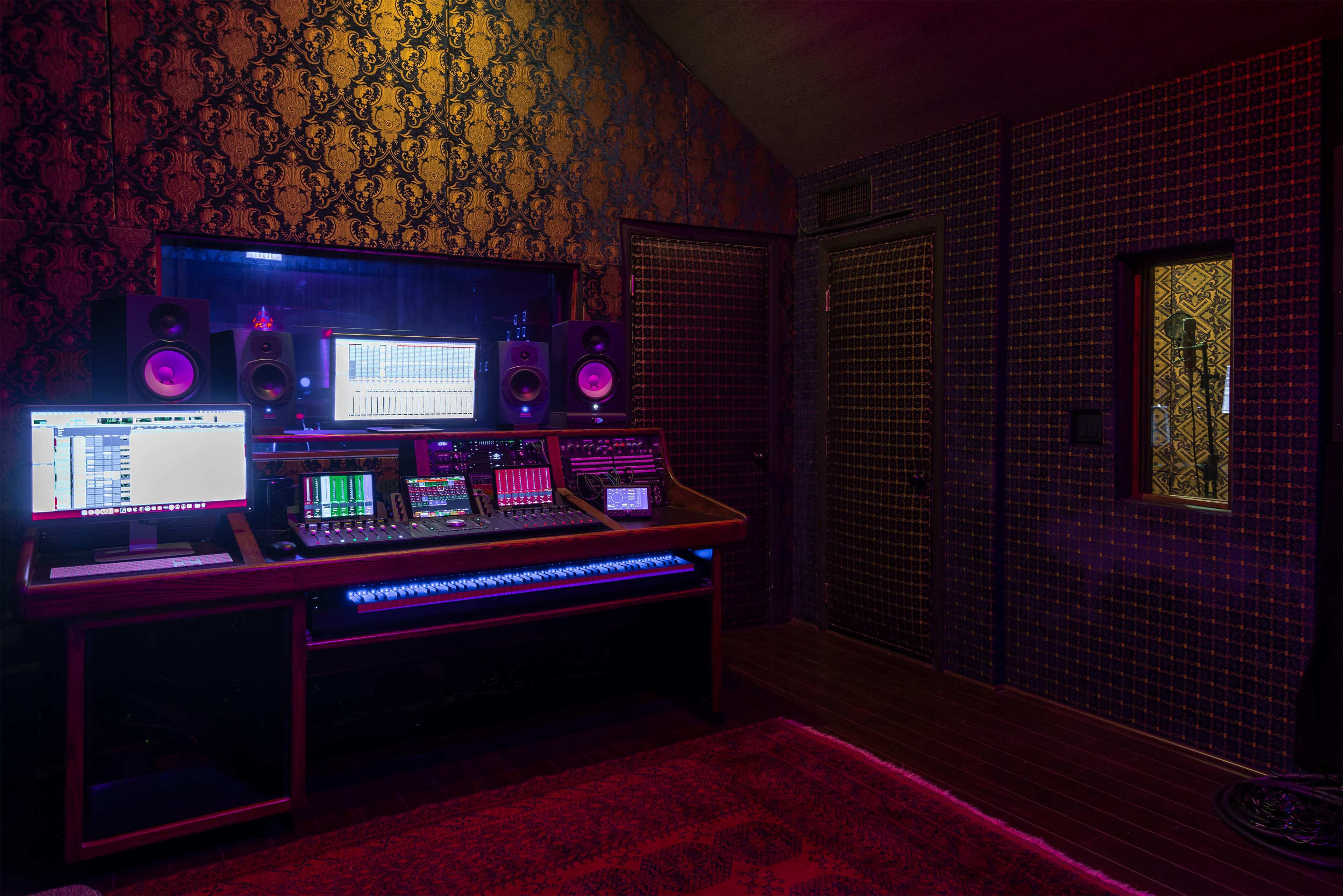 A dimly lit music studio with a large mixing console, multiple computer screens, and speakers arranged against a patterned wall.
