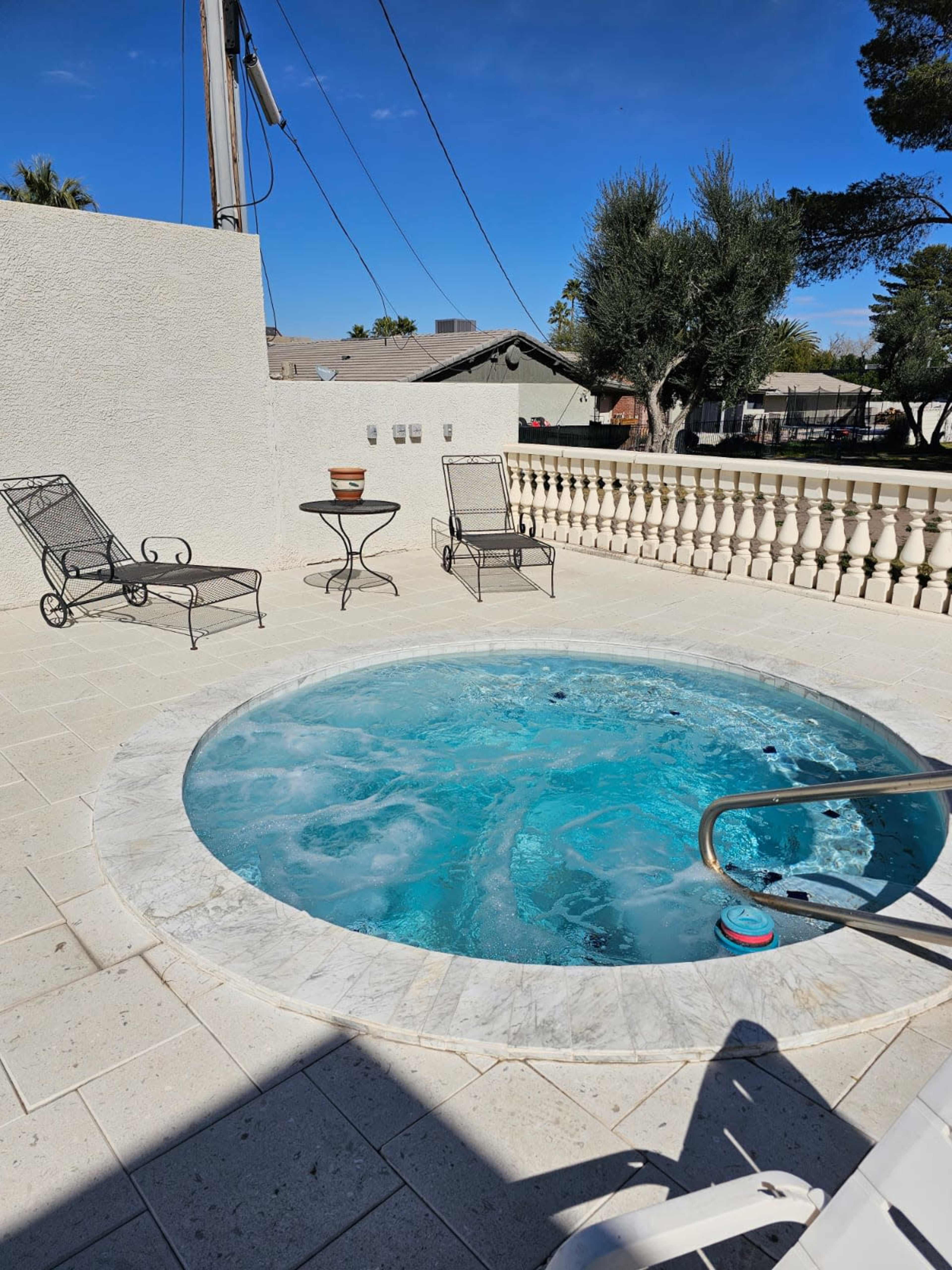 A hot tub on a patio surrounded by two lounge chairs and a table, with a fence and trees in the background.