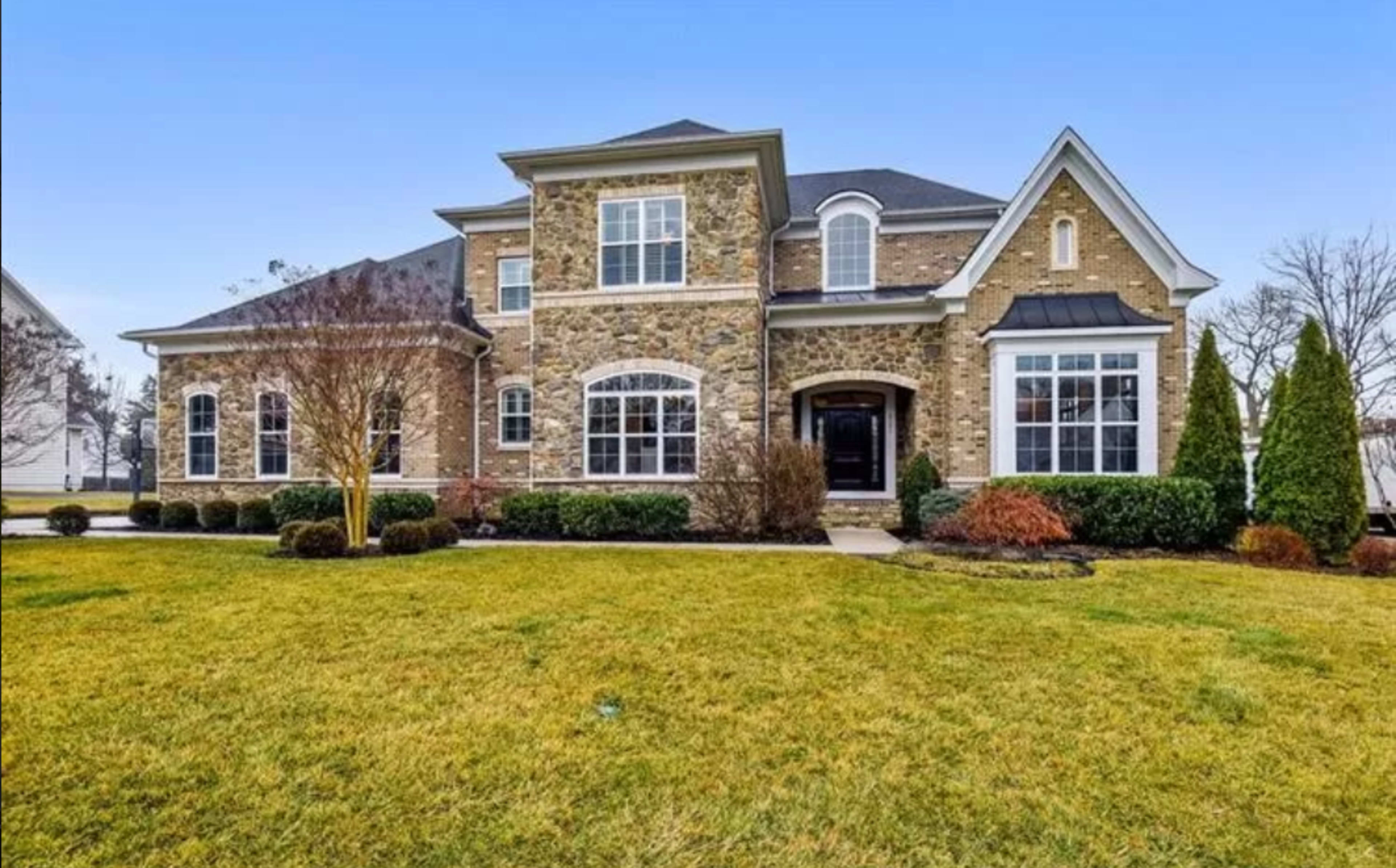 A large two-story house with stone and brick exterior is set on a landscaped lawn with neatly trimmed hedges.