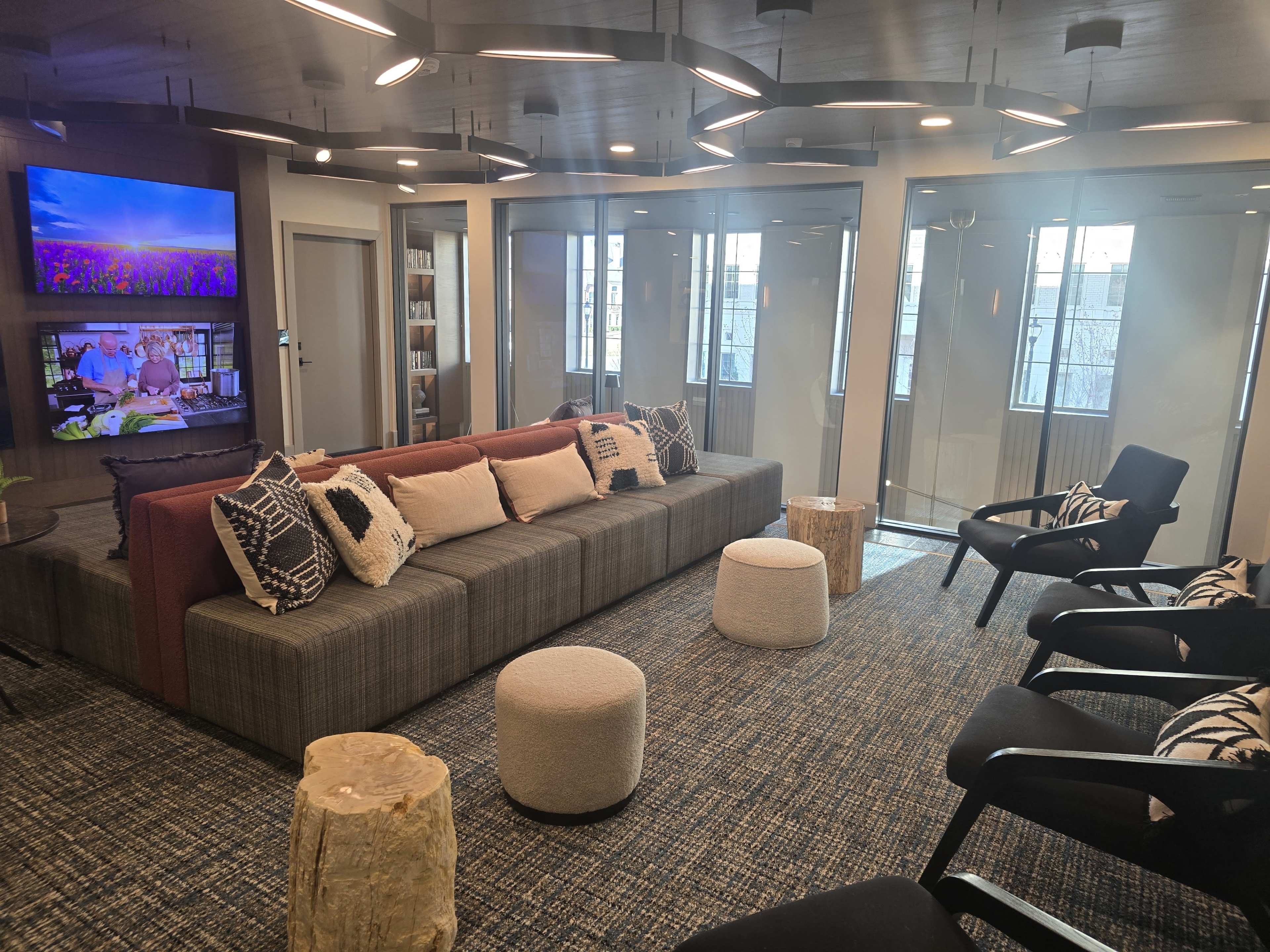 The image shows a modern lounge area with a sofa, several chairs, and wooden accents, featuring large windows and television screens on the walls.
