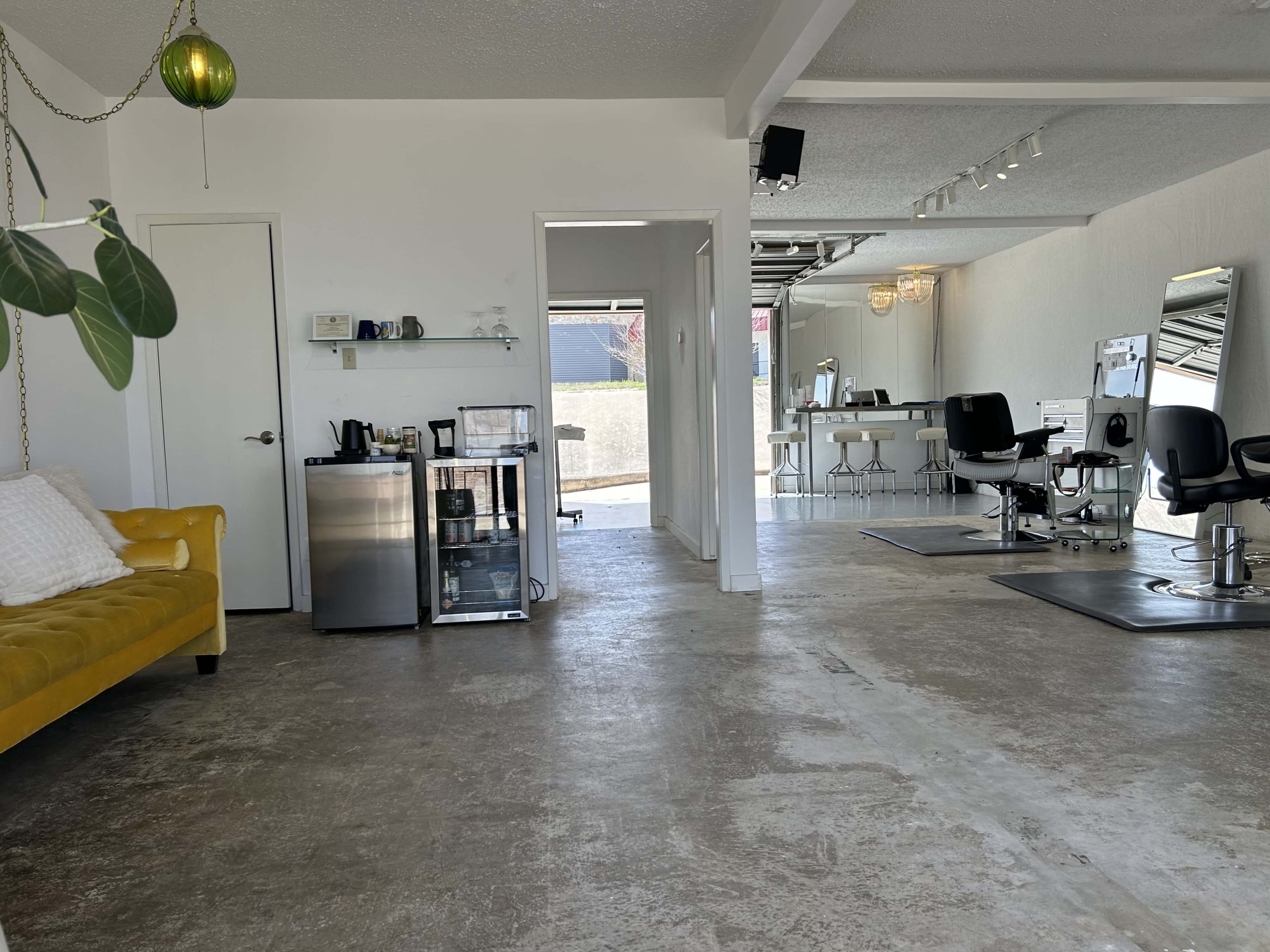 The image shows a spacious hair salon with a yellow couch, a beverage area, and multiple styling stations.