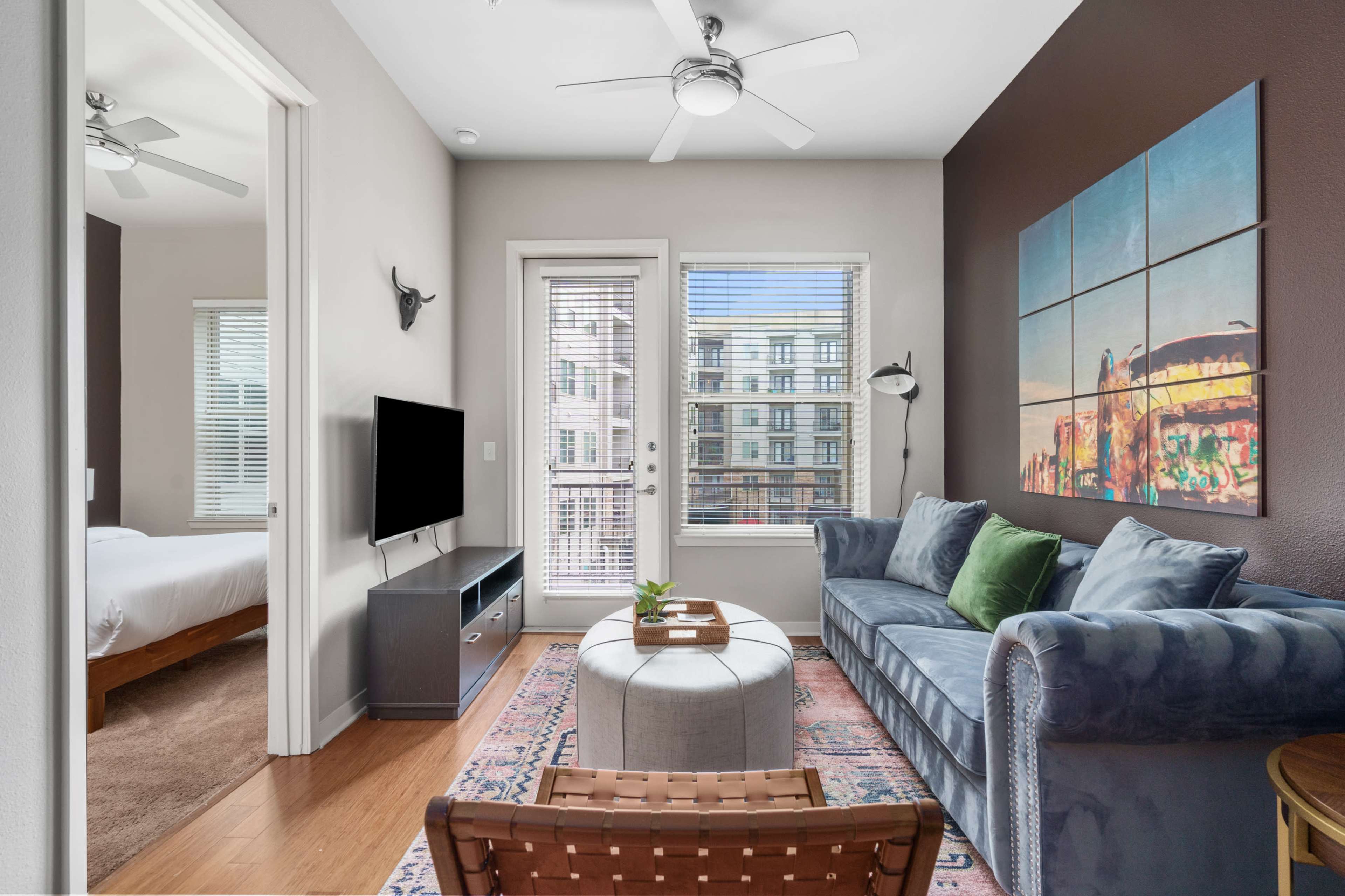 A living room features a blue couch, a round coffee table, a television, and a view of an outdoor balcony through glass doors.