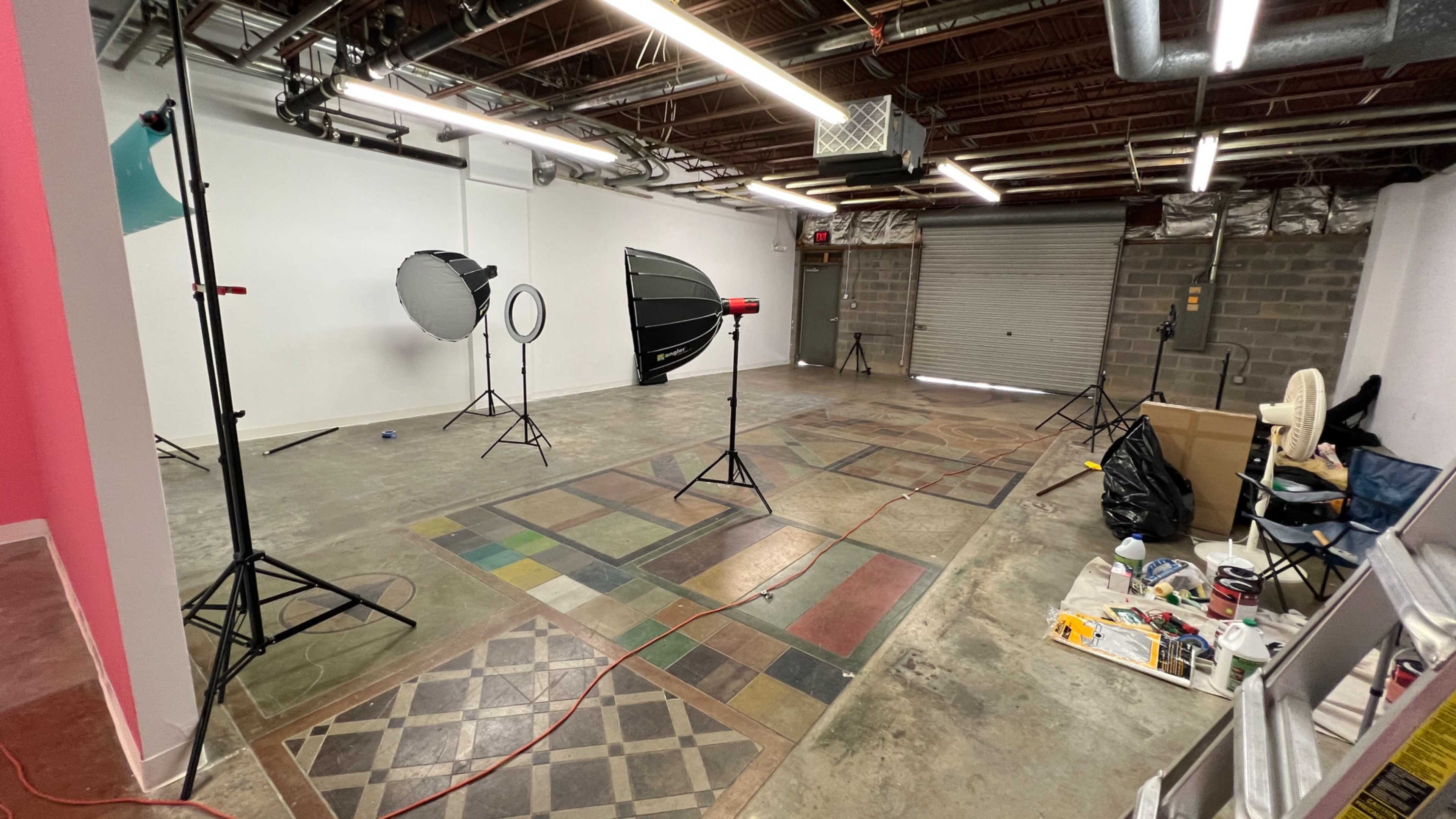 The image shows a photo studio setup featuring multiple lighting equipment, stands, and a partially cluttered floor in a spacious room with exposed ducts and a roll-up garage door.