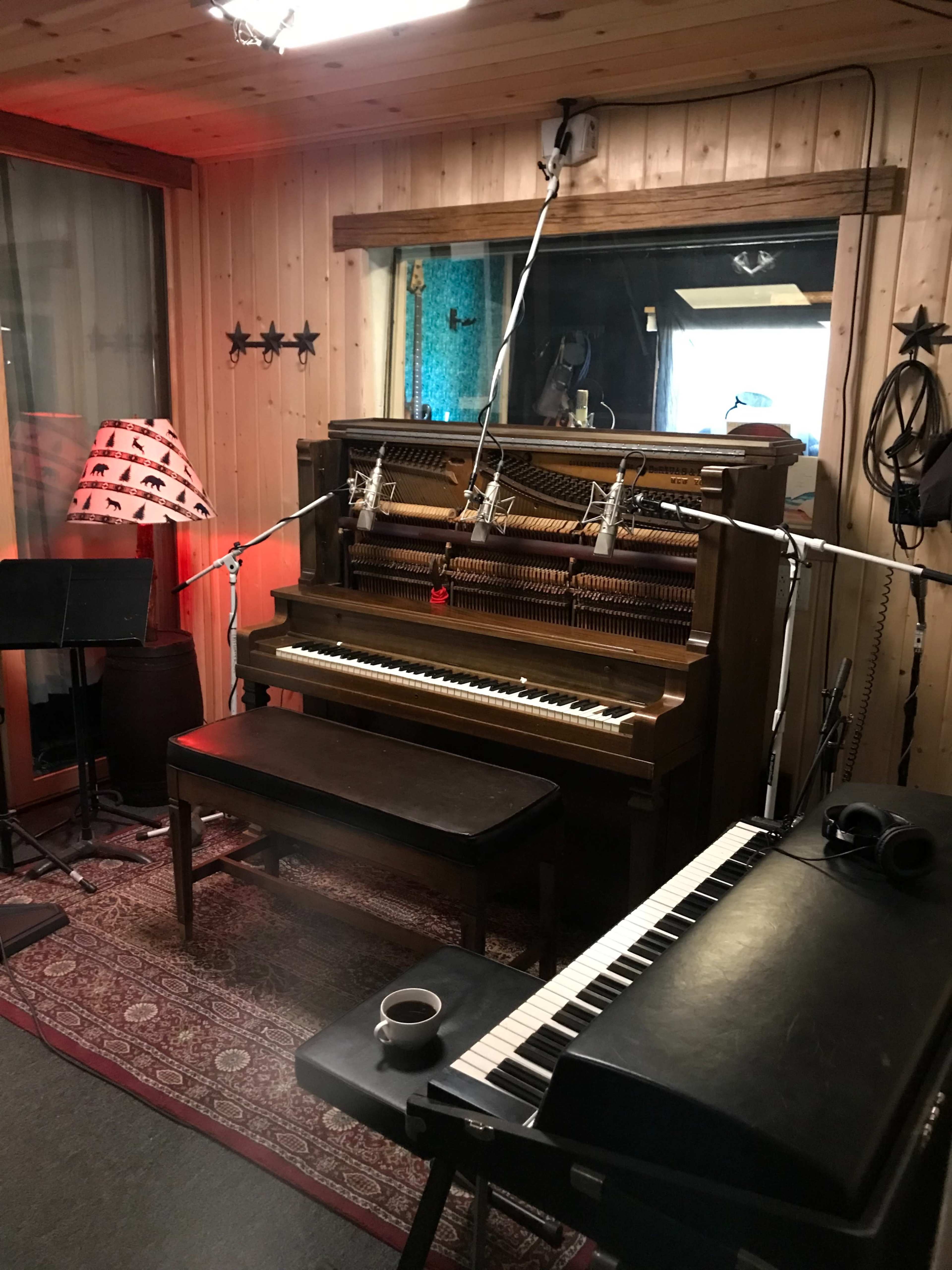 A wooden piano with a matching bench is set up in a recording studio, surrounded by microphones and a coffee cup on a keyboard.