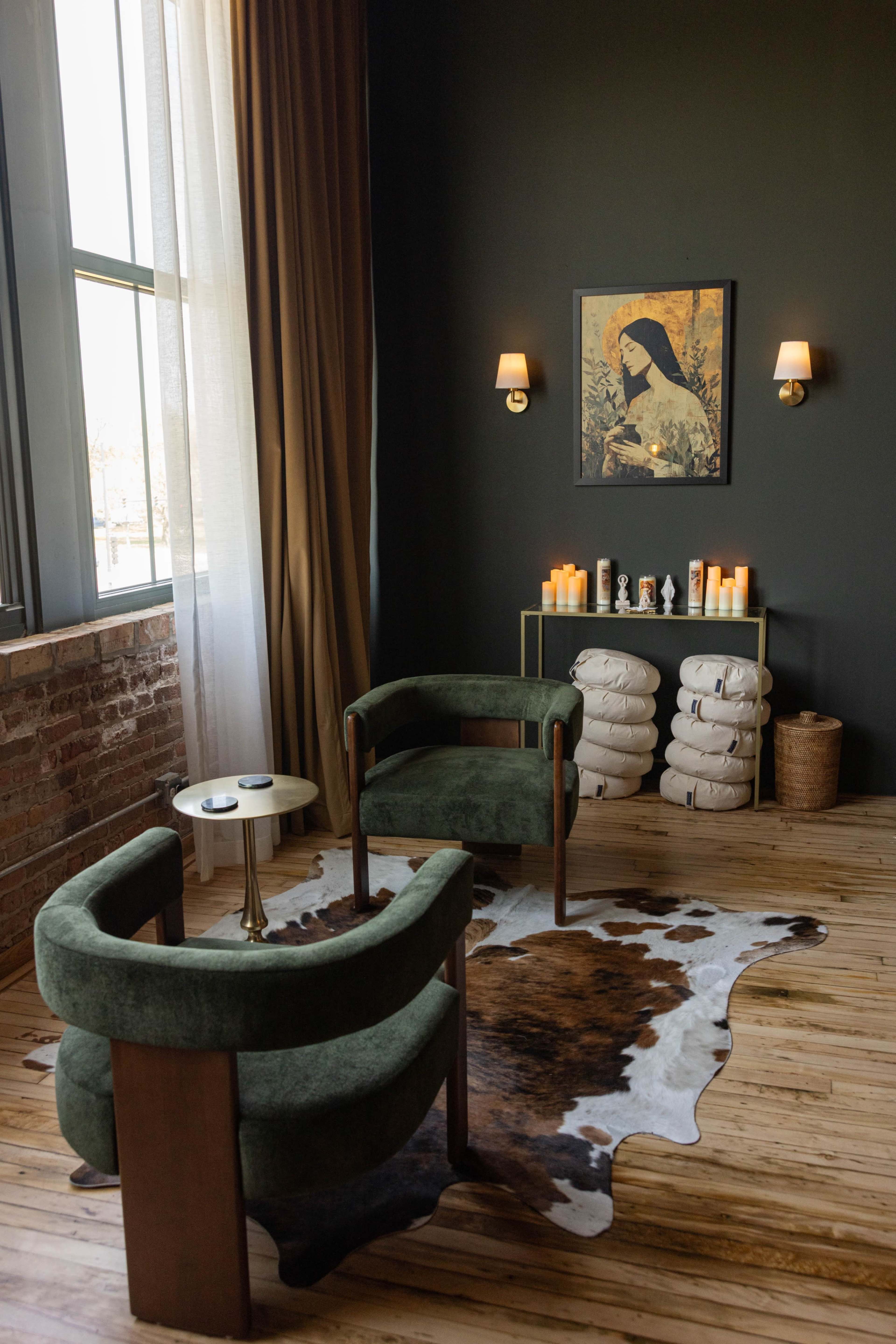 The room features two green armchairs facing a small table, with a cowhide rug on the wooden floor and shelves displaying candles and towels against a dark wall.