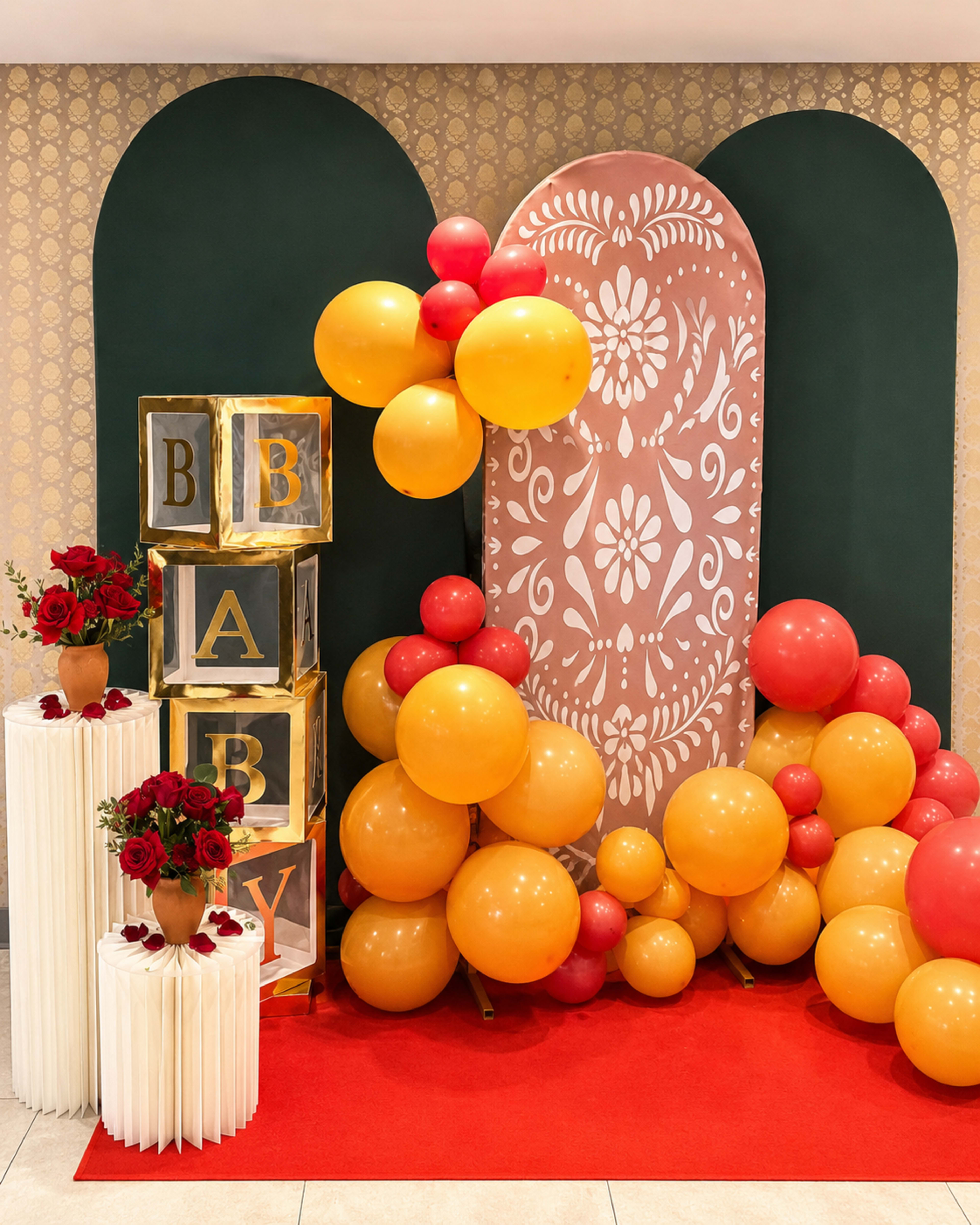 The image shows a decorative setup featuring gold and red balloons, floral arrangements, and large gold letter blocks spelling "BABY" against a patterned backdrop.