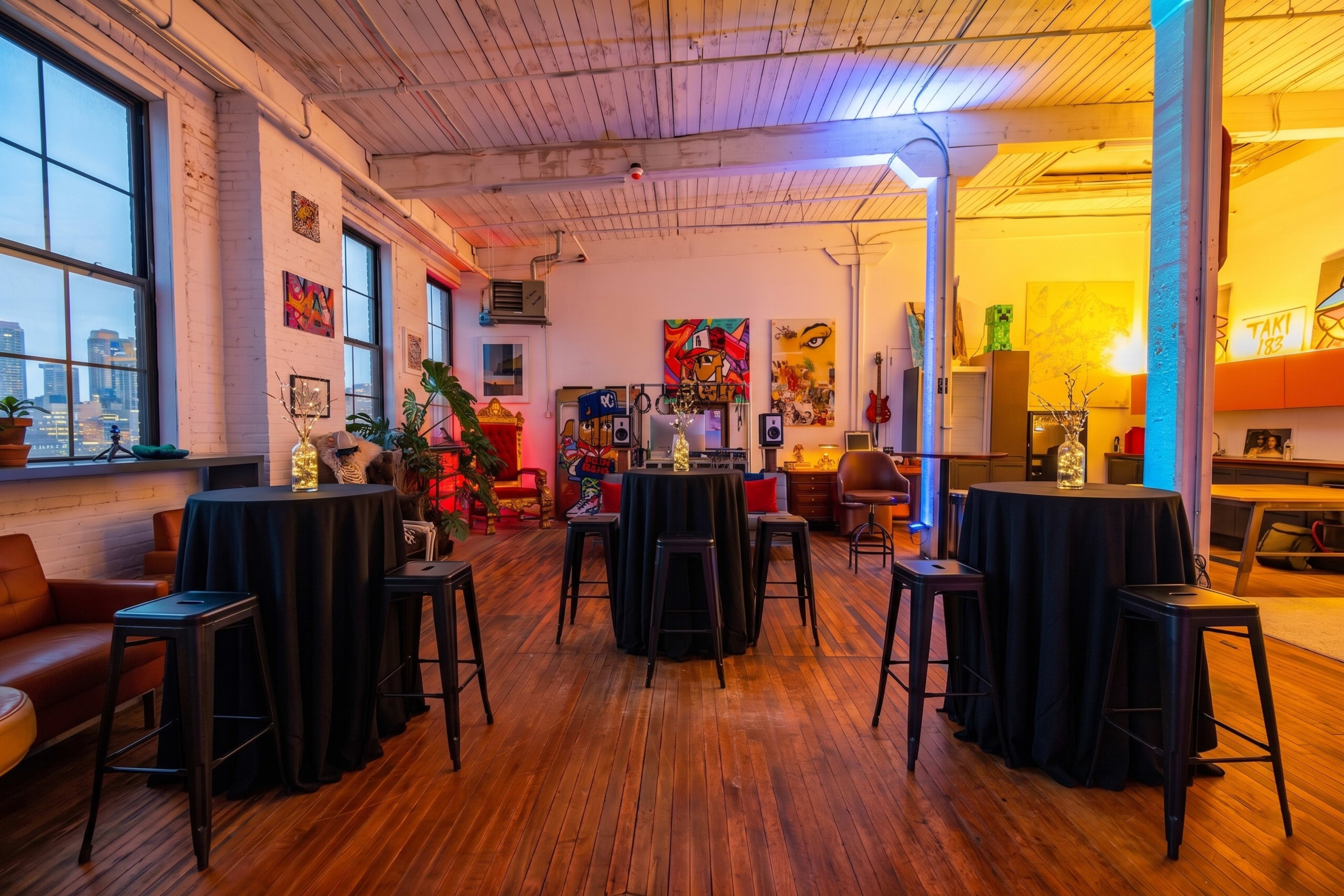 The image shows a well-lit, eclectic space with several tall tables covered in black cloth, surrounded by colorful artwork and furniture pieces.