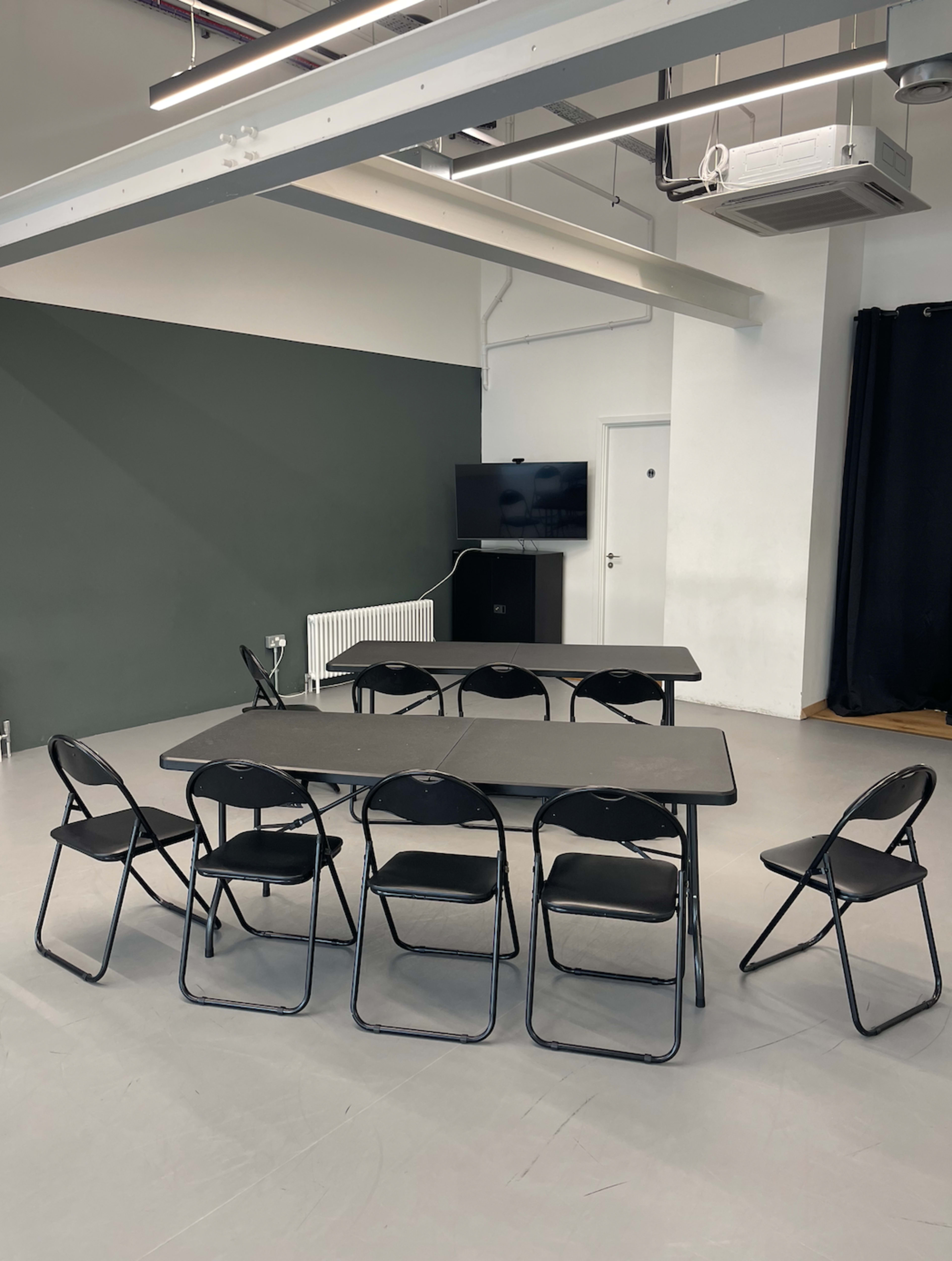 A rectangular table is surrounded by six folding chairs in a modern room with a dark green wall and a television on a stand.