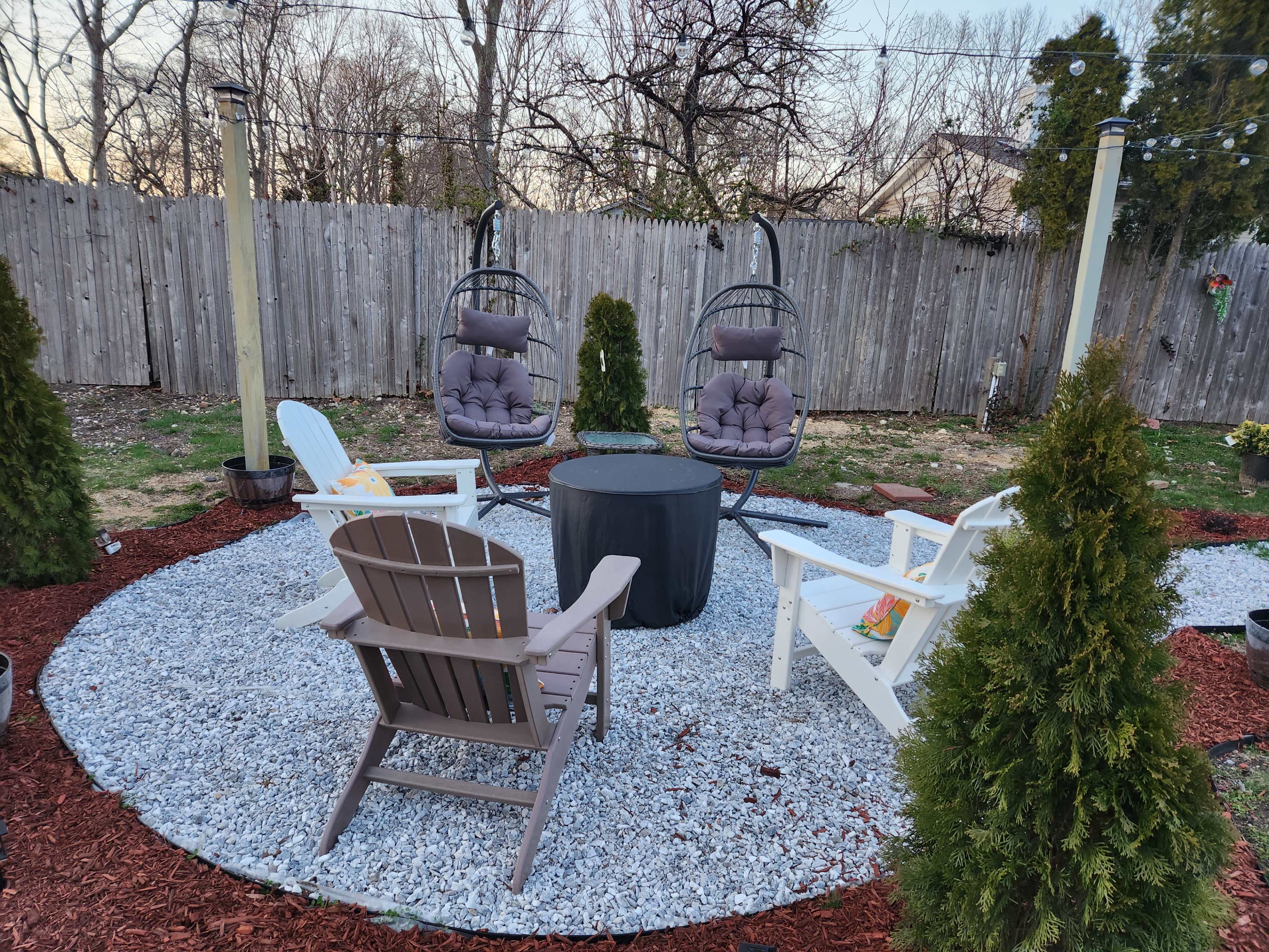 A small outdoor seating area featuring two hanging chairs and four additional chairs arranged around a circular gravel platform, surrounded by decorative shrubs and a wooden fence.