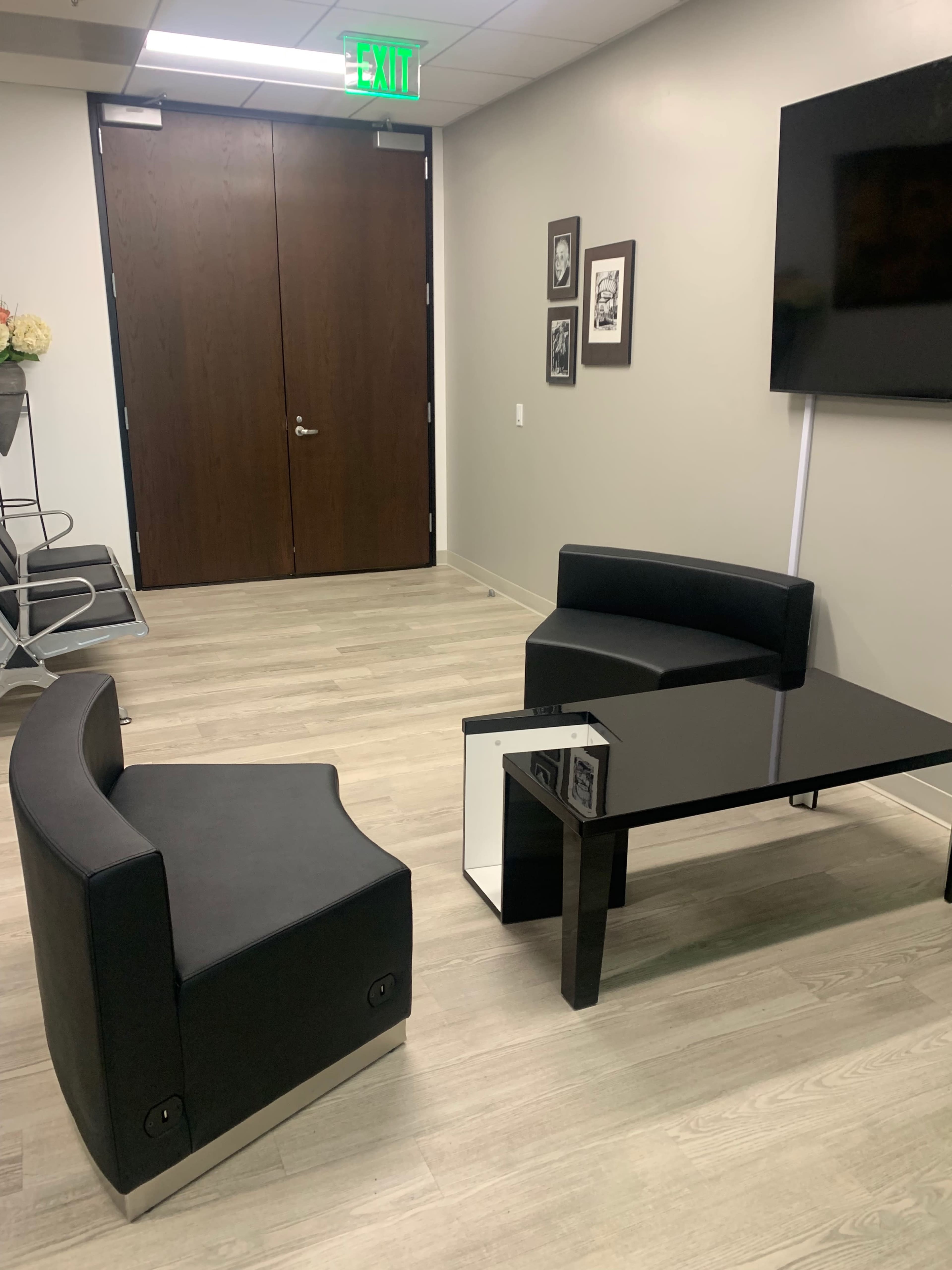 The image shows a minimalist office waiting area with two black curved chairs and a black glass coffee table, situated near a wooden door and a television.