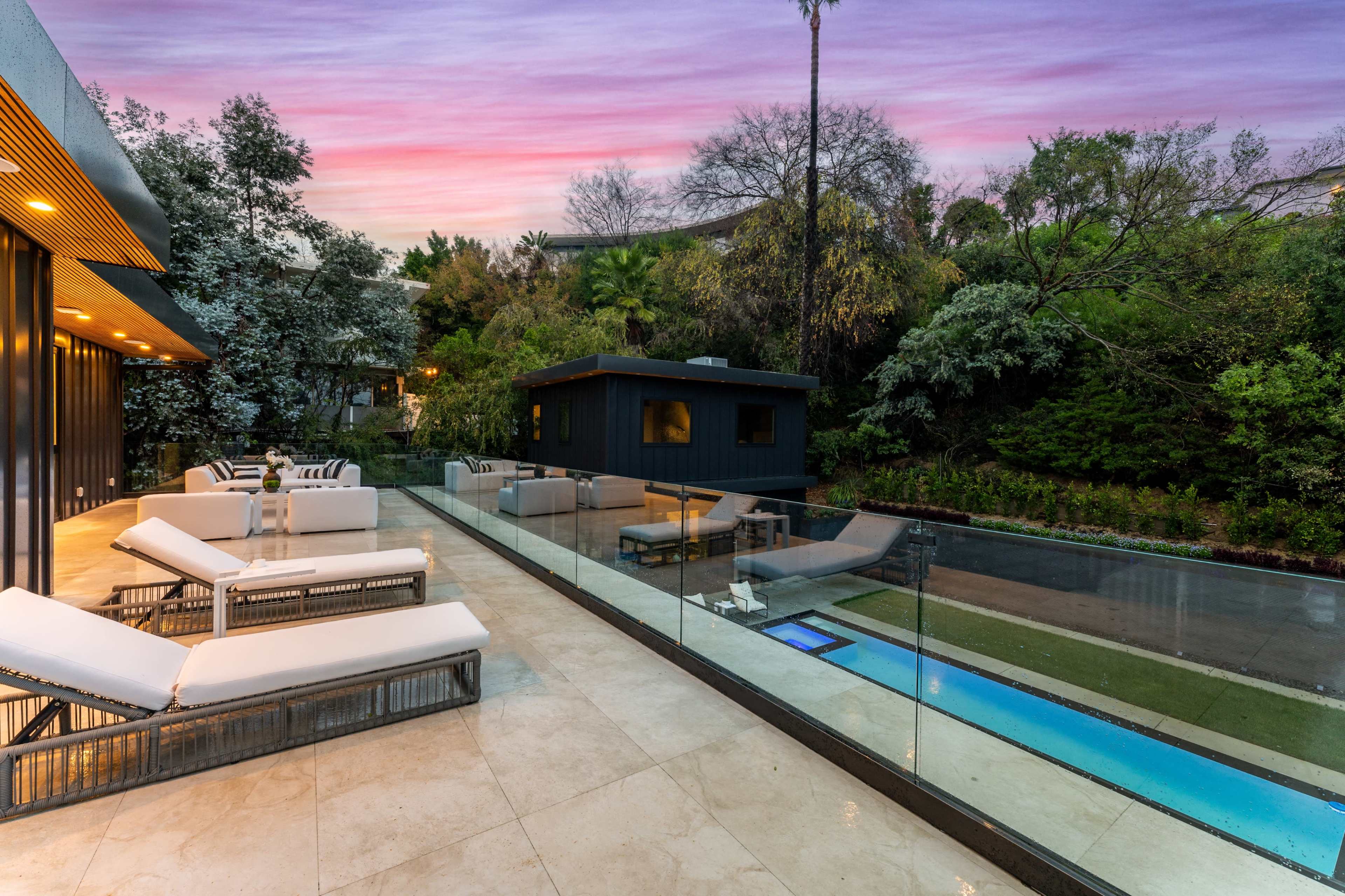 The image shows a modern outdoor lounge area with white sun loungers beside a narrow swimming pool and a backdrop of lush greenery under a colorful sky at dusk.
