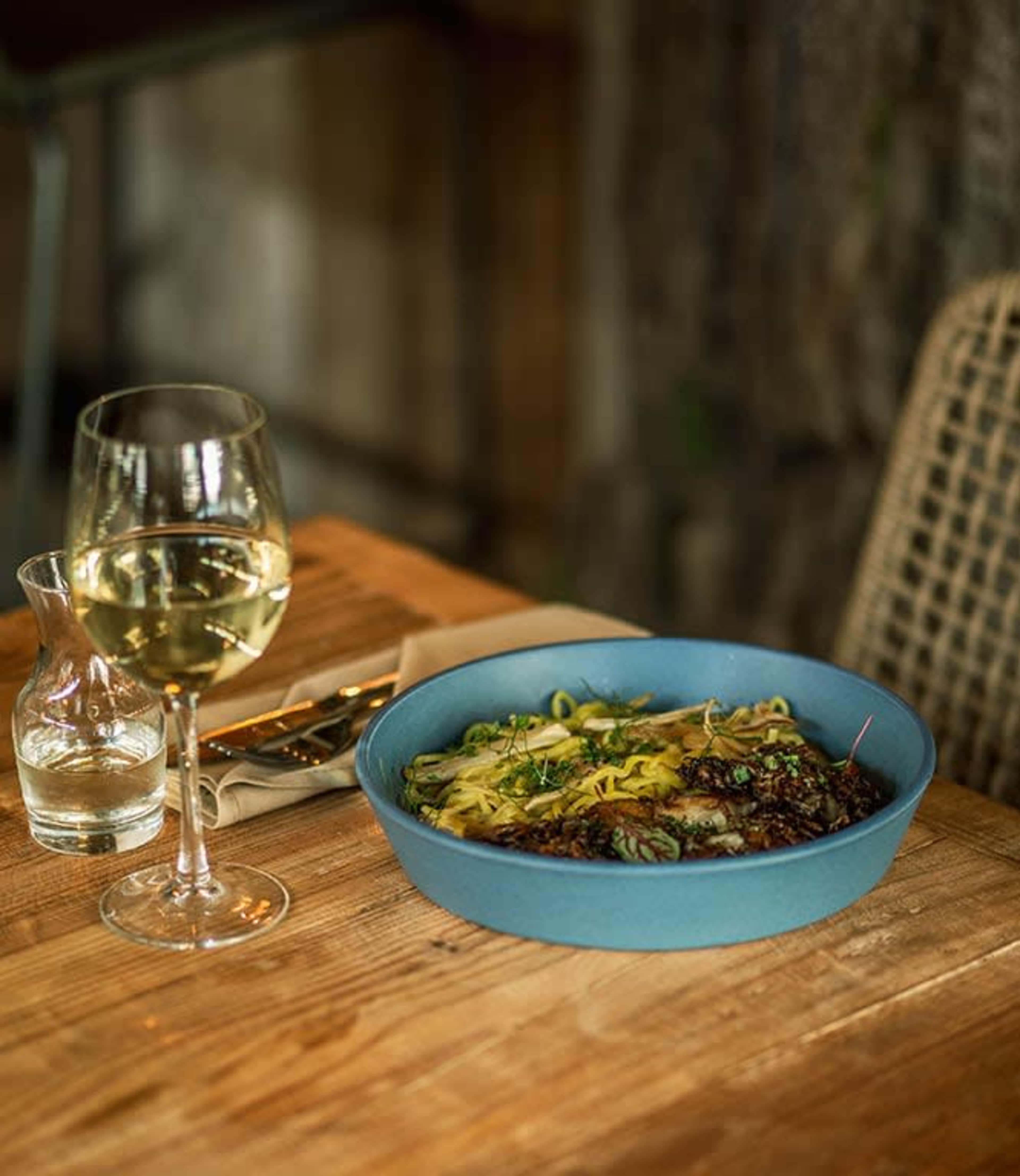 A blue bowl filled with a colorful dish of pasta and garnishes sits on a wooden table next to a glass of white wine and a small carafe of water.