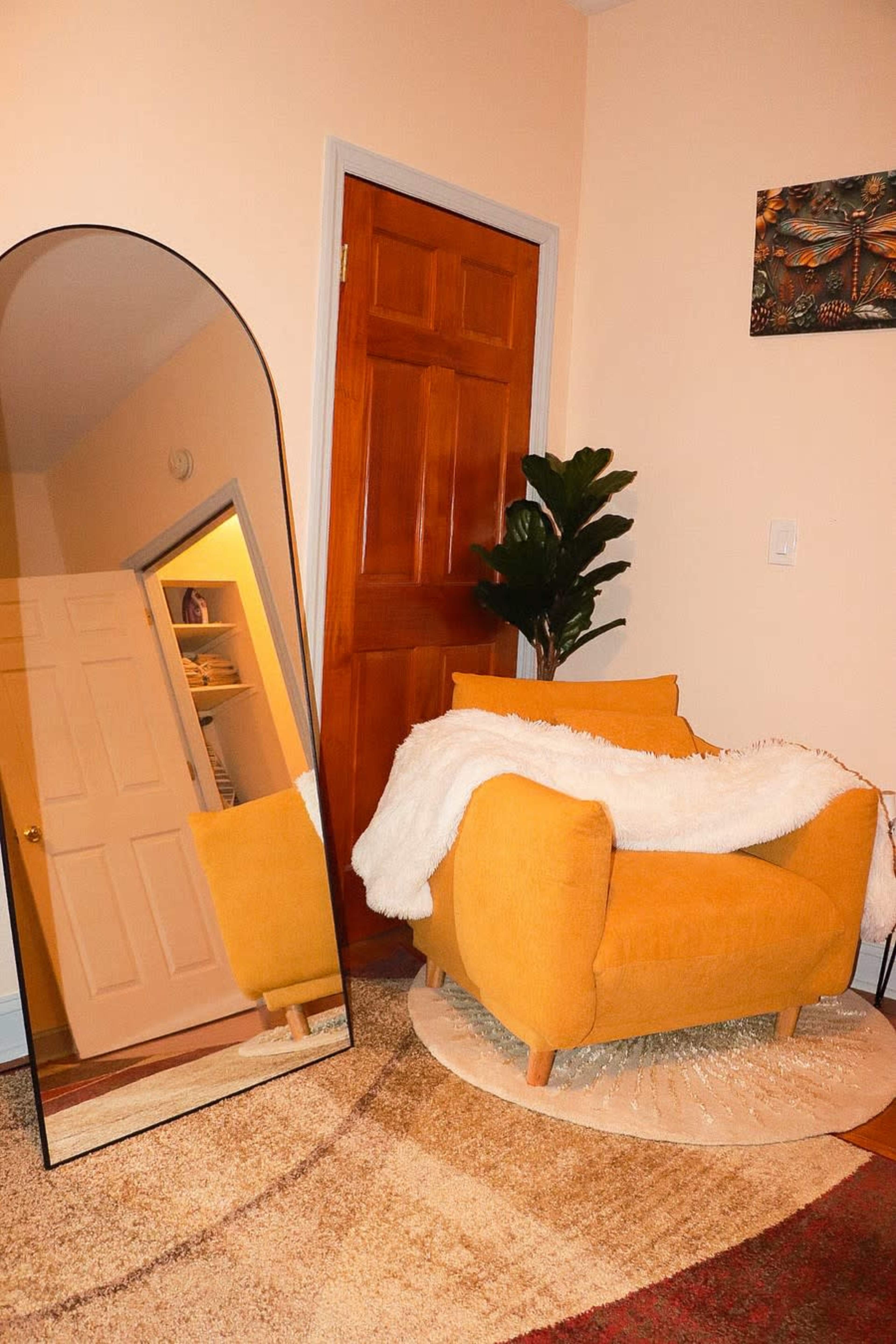 The image shows a cozy room featuring a large, arched mirror reflecting a bright yellow armchair draped with a white throw, alongside a potted plant and a closed wooden door.