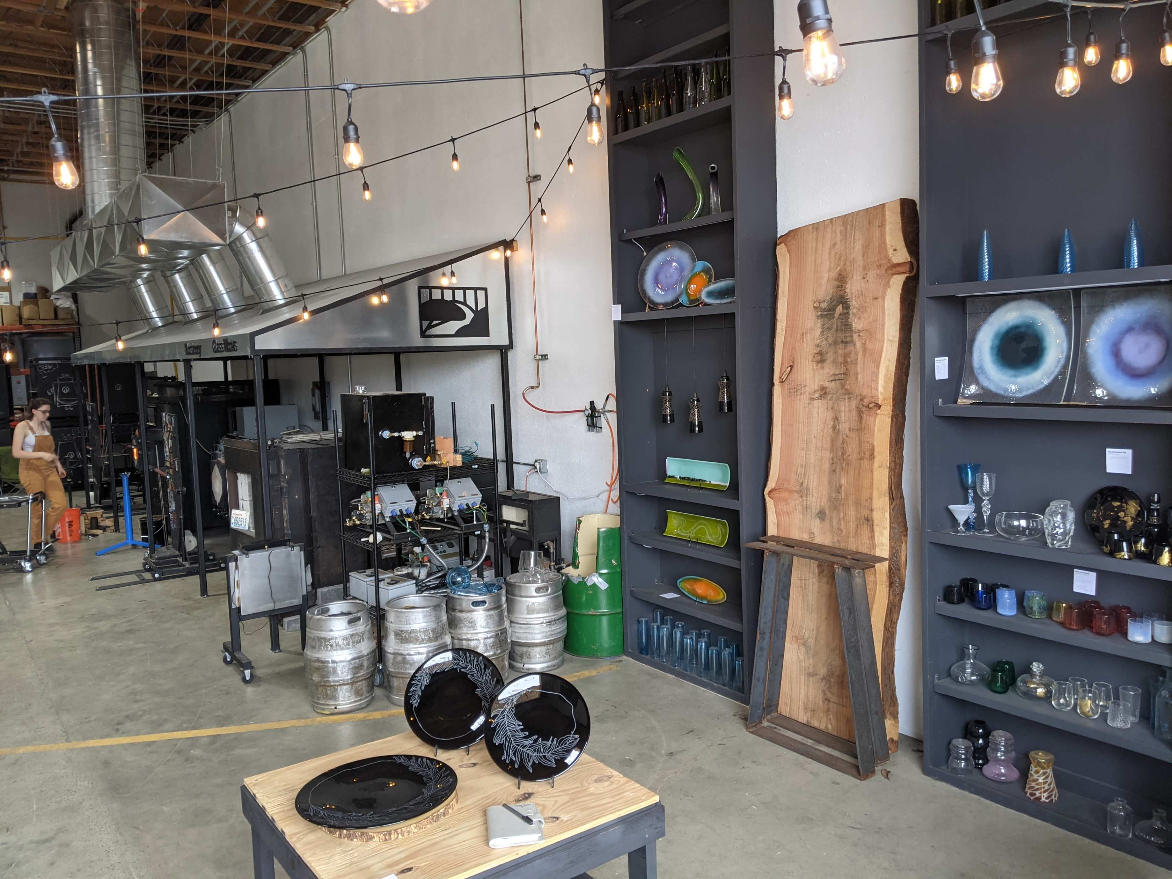 An industrial-style glassblowing studio with various glass artworks displayed on shelves, a work area with equipment, and a wooden table in the foreground.