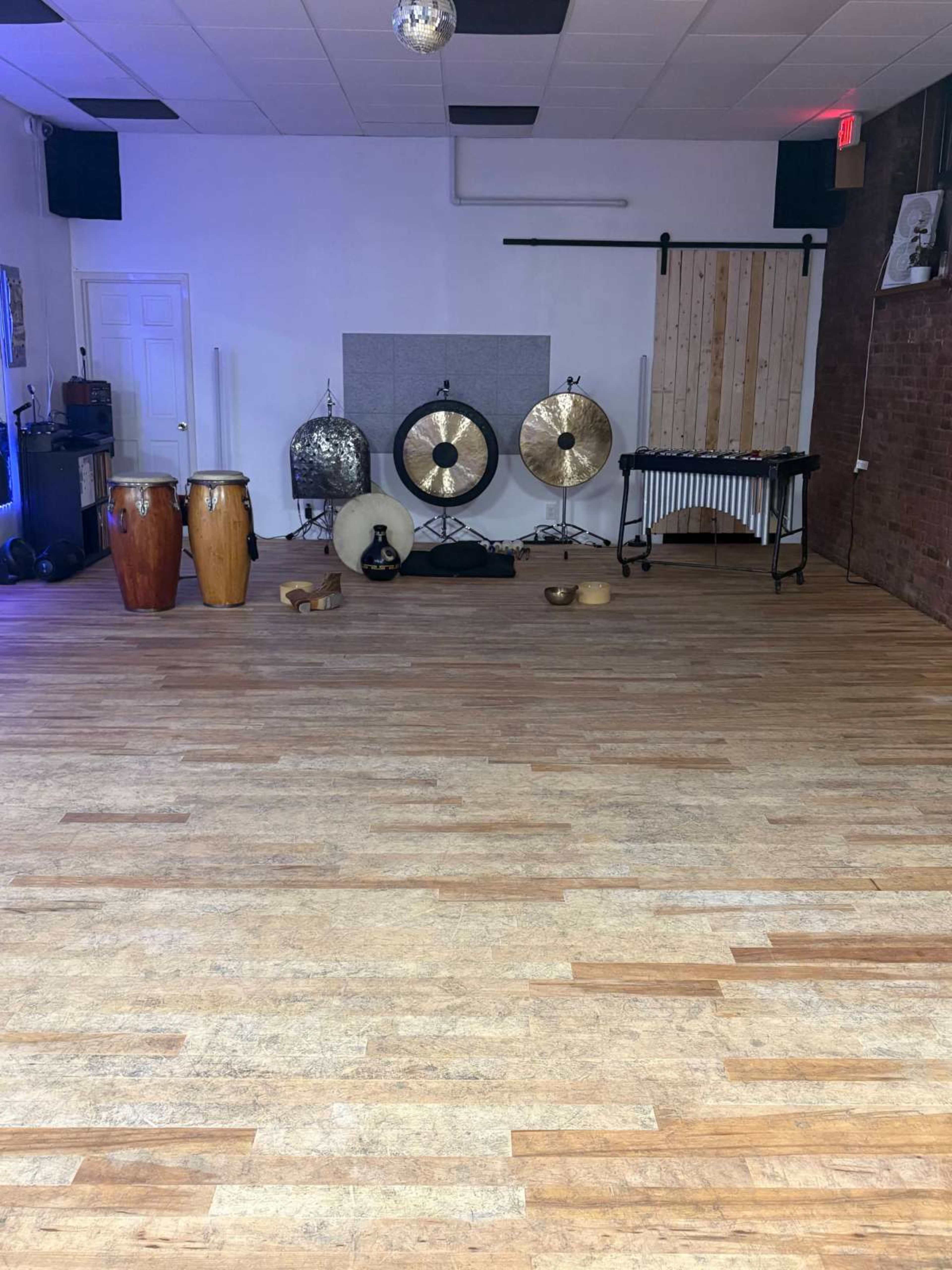 The image shows a spacious room with wooden flooring, featuring various musical instruments, including congas, gongs, cymbals, and a vibraphone, arranged neatly in one area.