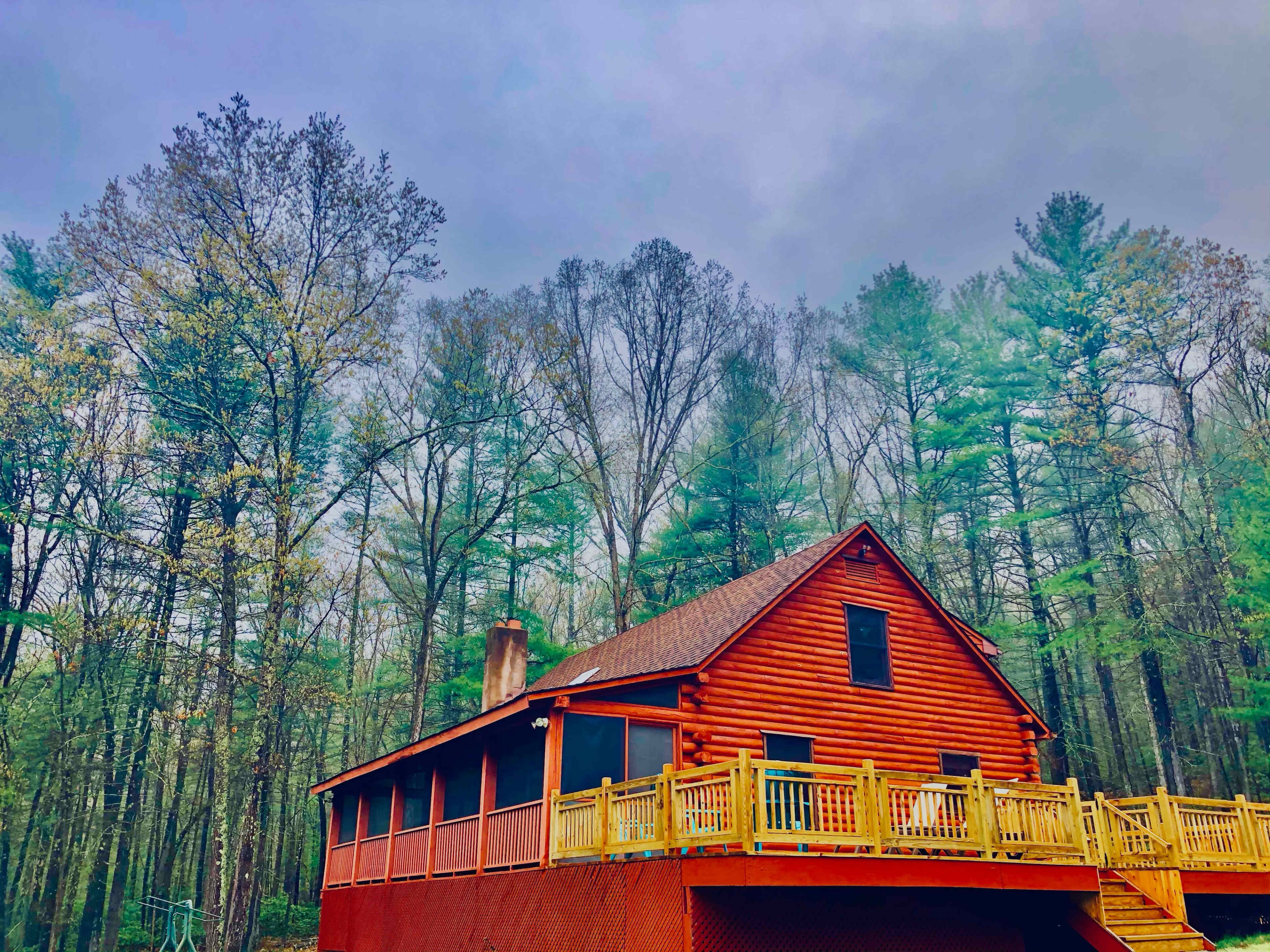A red wooden cabin with a deck is surrounded by tall trees under a cloudy sky.