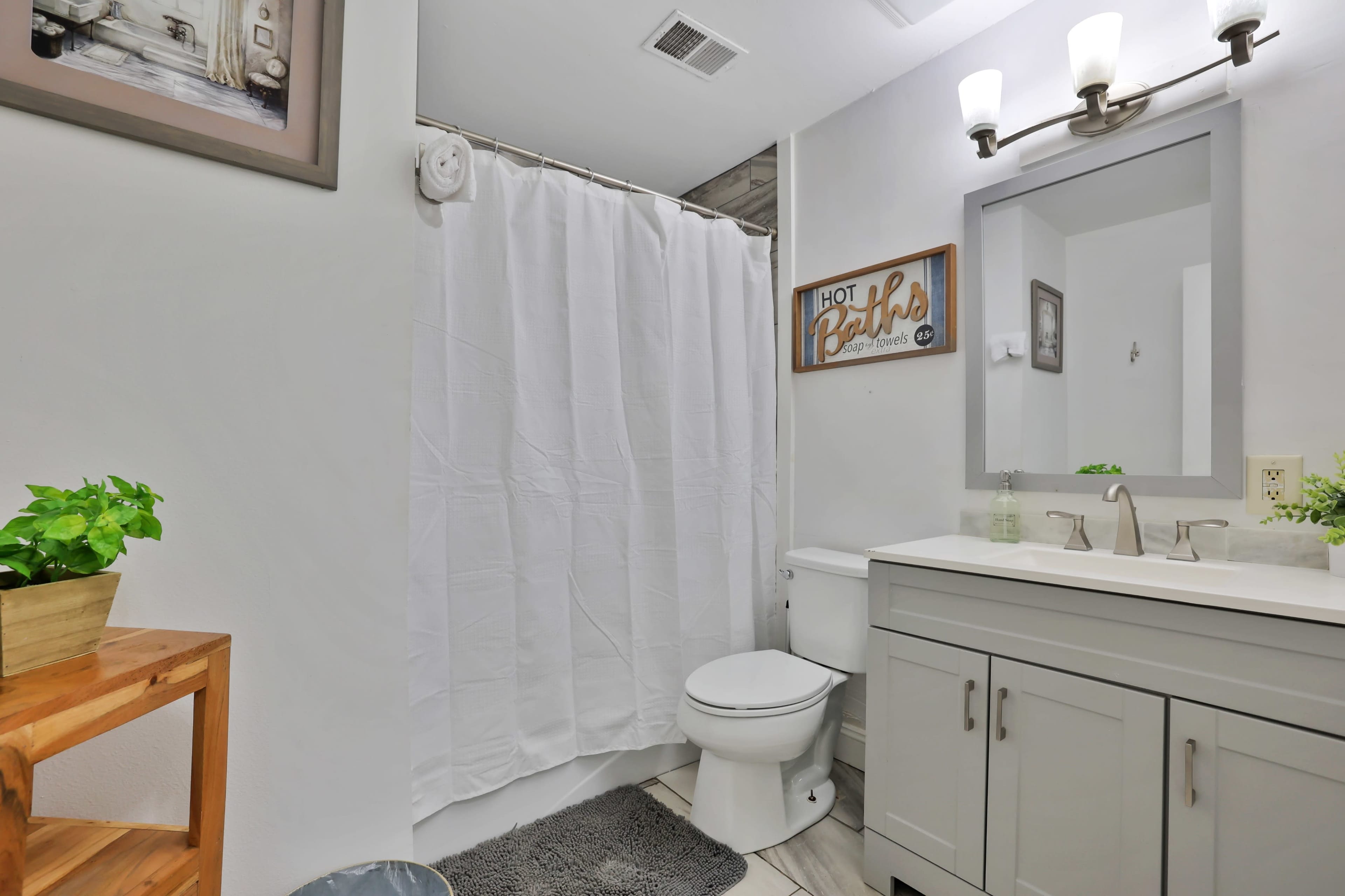 A clean bathroom features a white shower curtain, a gray vanity with a mirrored cabinet, a toilet, and a small shelf with a potted plant.