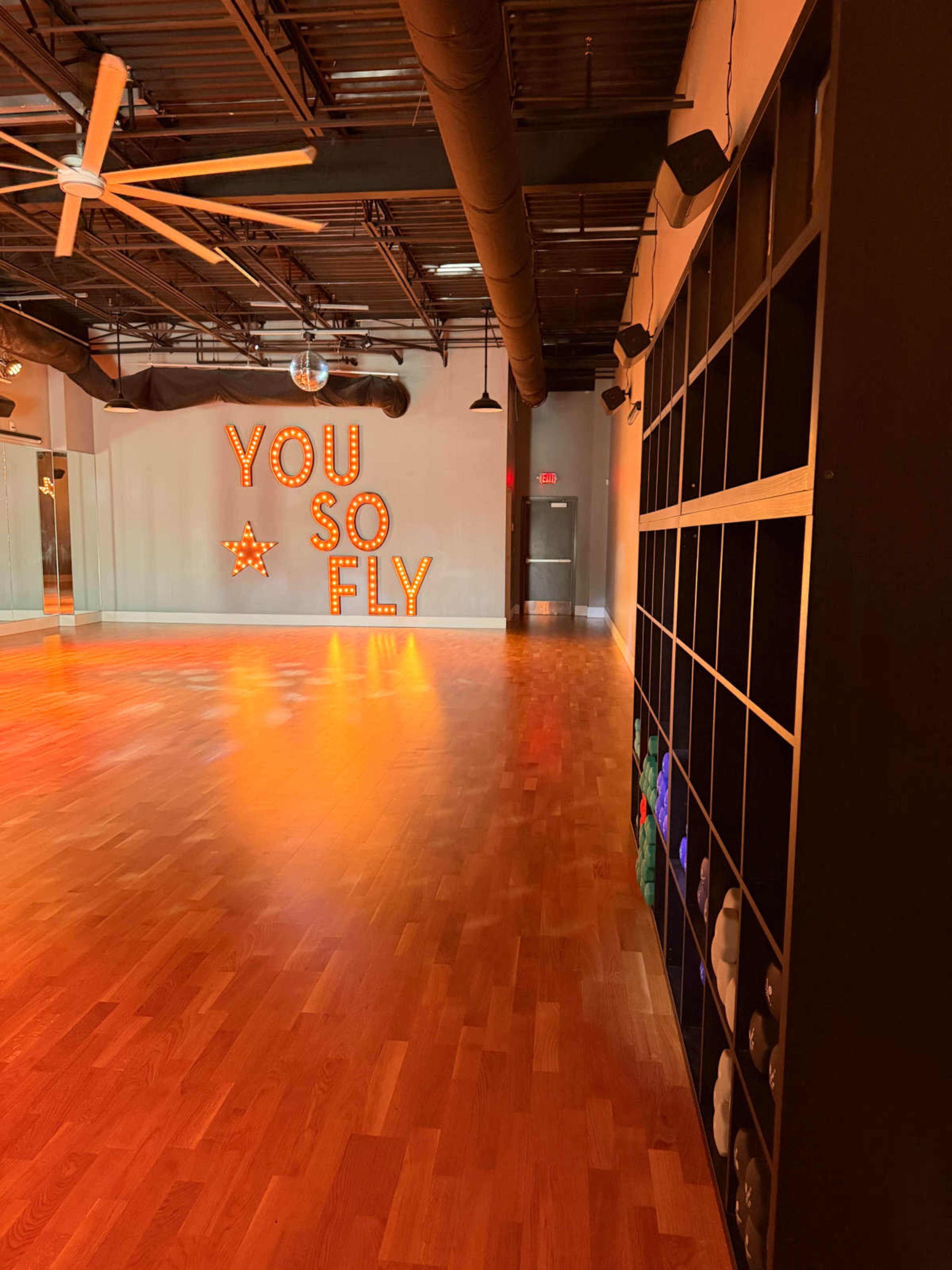 The image shows a dance studio with wooden floors, wall-mounted shelving, and illuminated letters spelling "YOU SO FLY" against a brightly lit backdrop.
