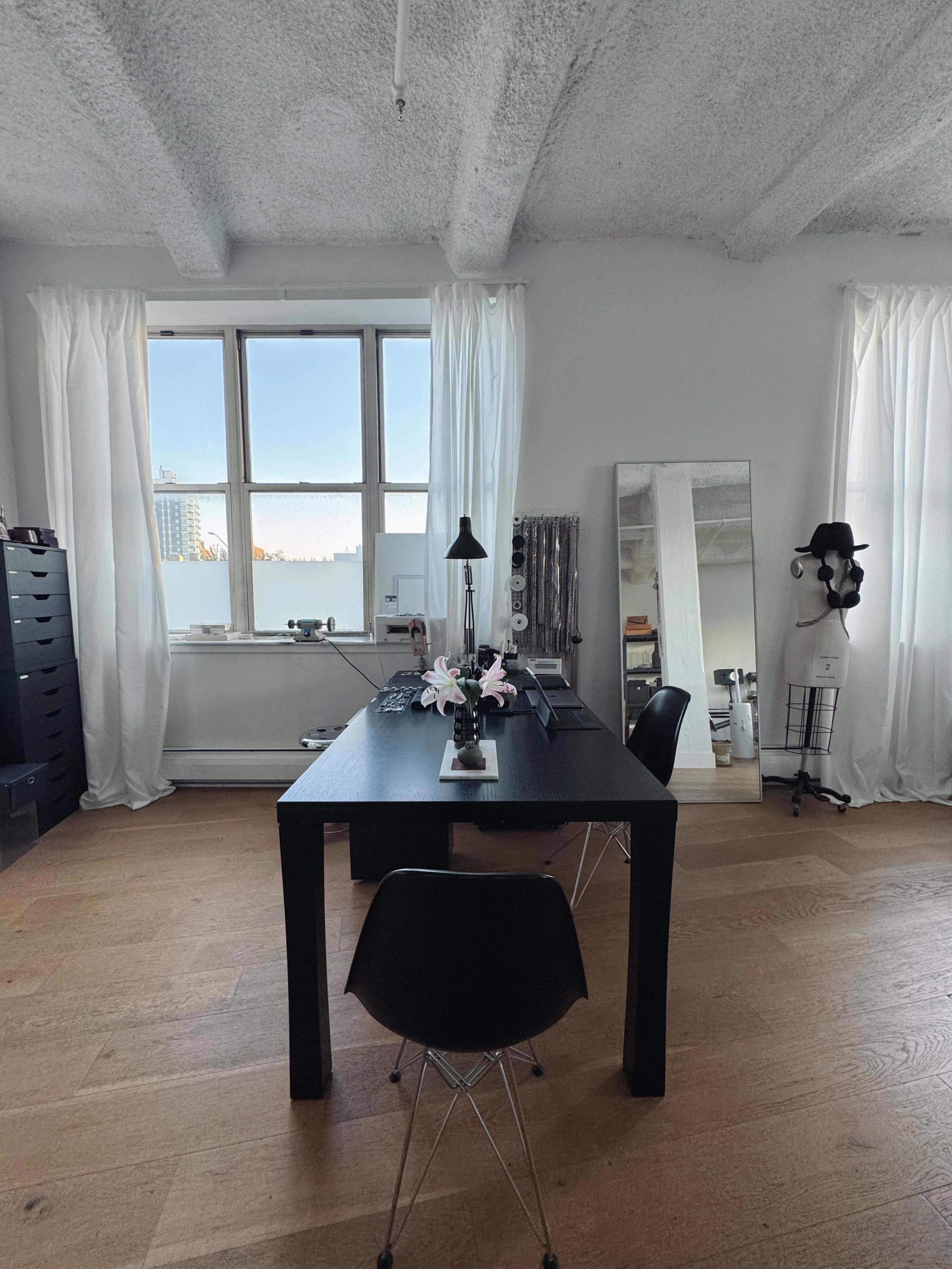 The image shows a minimalist office space featuring a black desk and chair, a large mirror, and large windows with white curtains.
