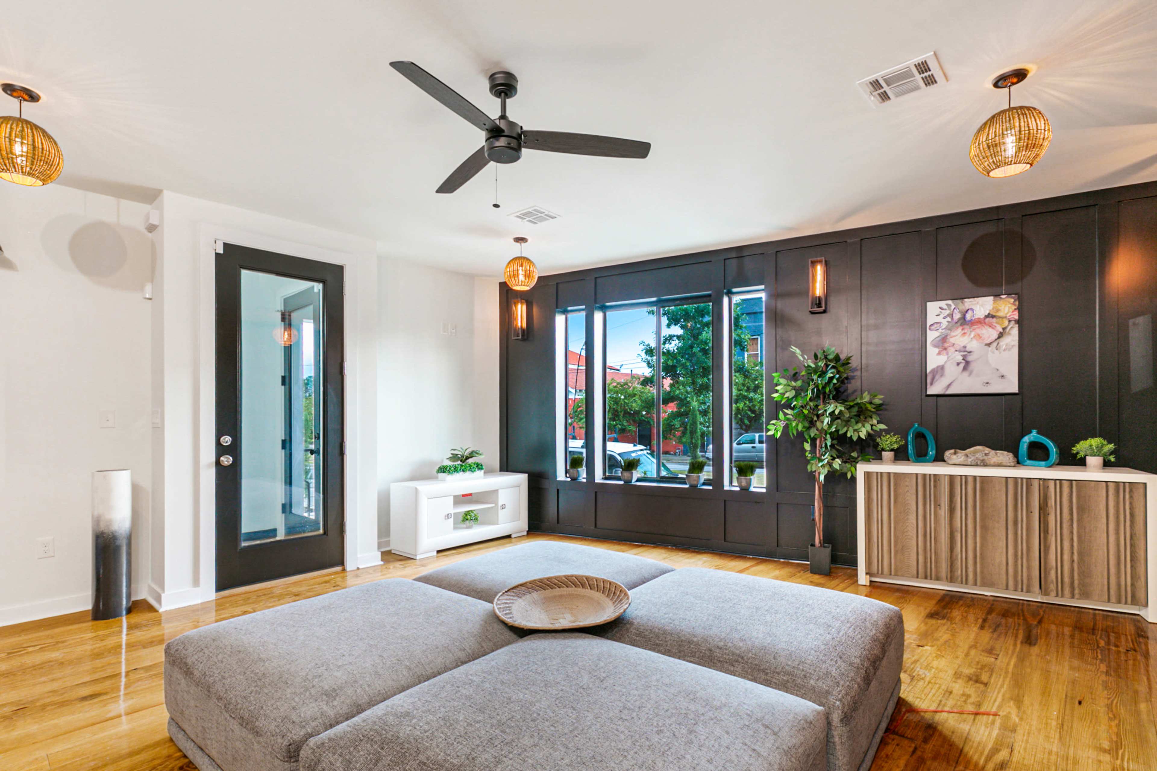 A spacious living room features a large gray ottoman, a ceiling fan, and large windows with greenery visible outside.