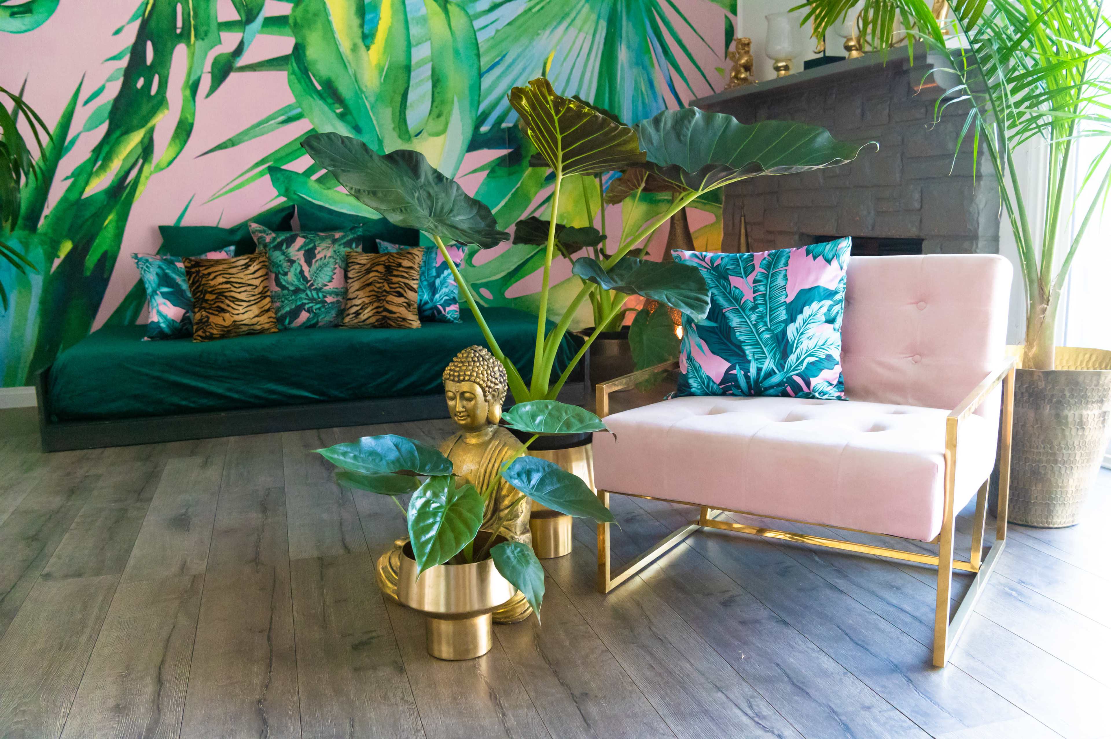 A pink chair with gold accents sits beside a gold Buddha statue and a plant, in a room adorned with leafy green wallpaper and decorative pillows.